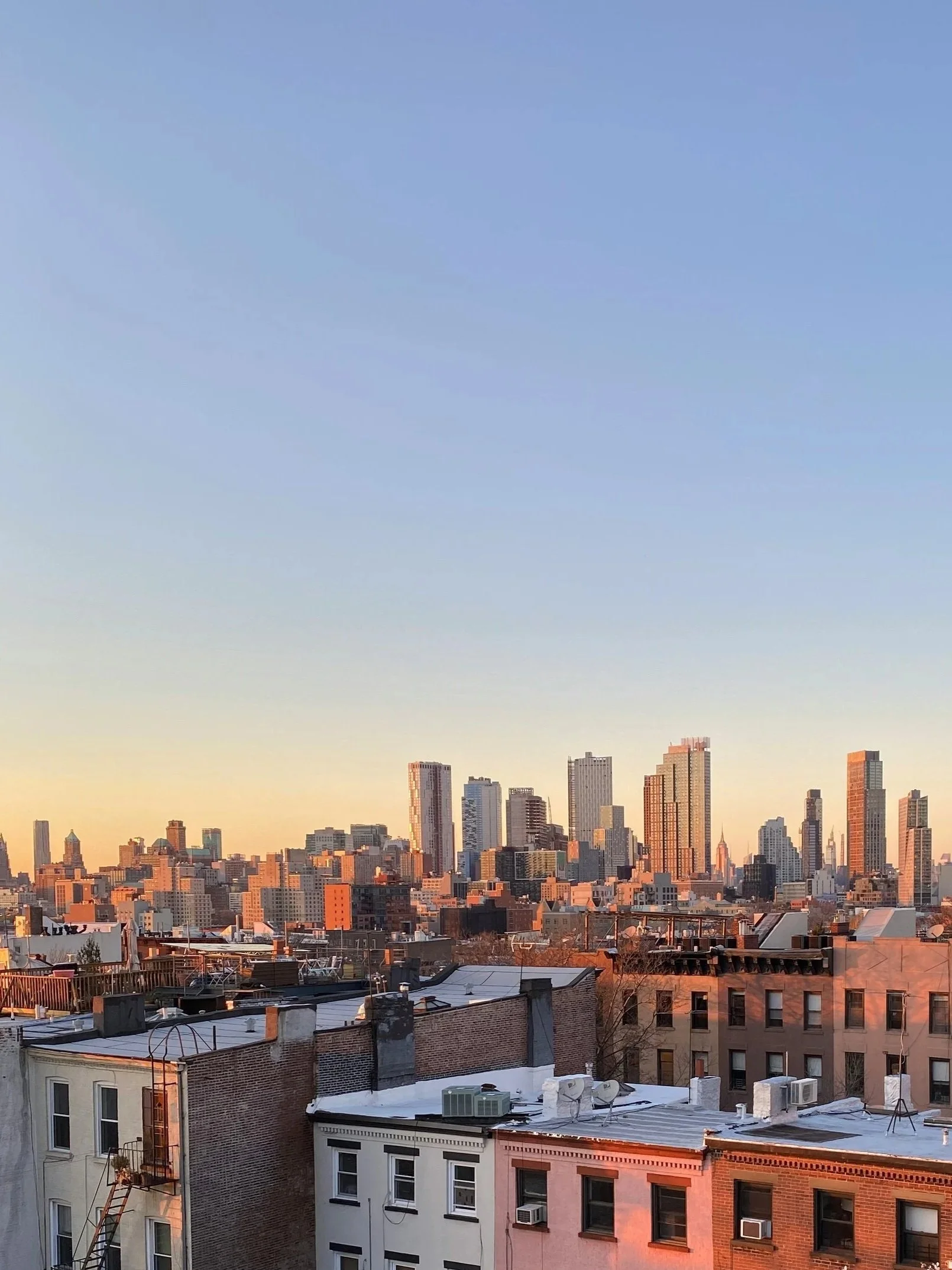 The image shows a cityscape at sunset with a clear sky. Tall buildings and skyscrapers are visible in the background, with lower-rise residential buildings in the foreground.