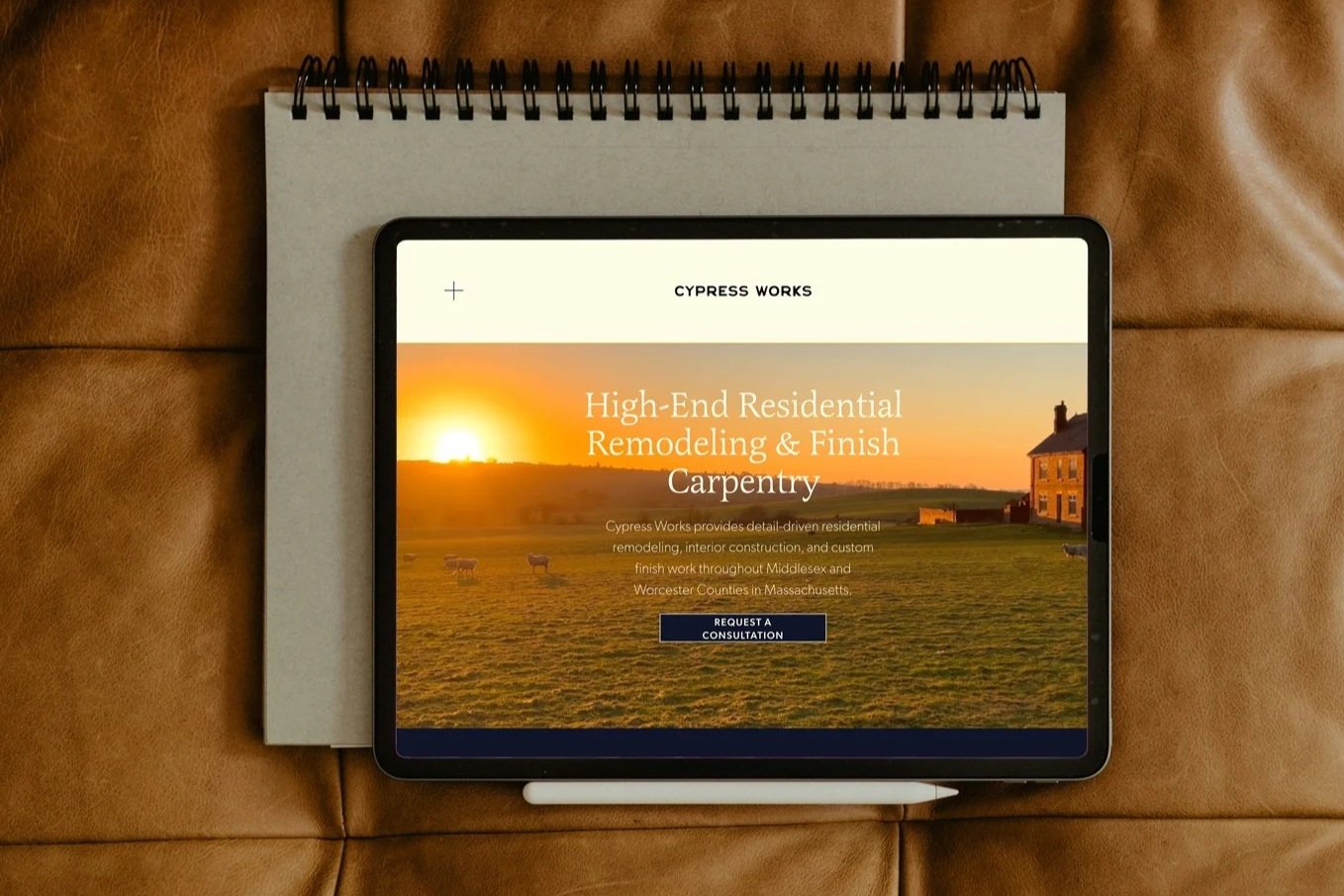 An electronic tablet displaying a webpage about high-end residential remodeling and carpentry, with a sunset and rural landscape background. The tablet is placed on a white notepad on a brown leather surface.