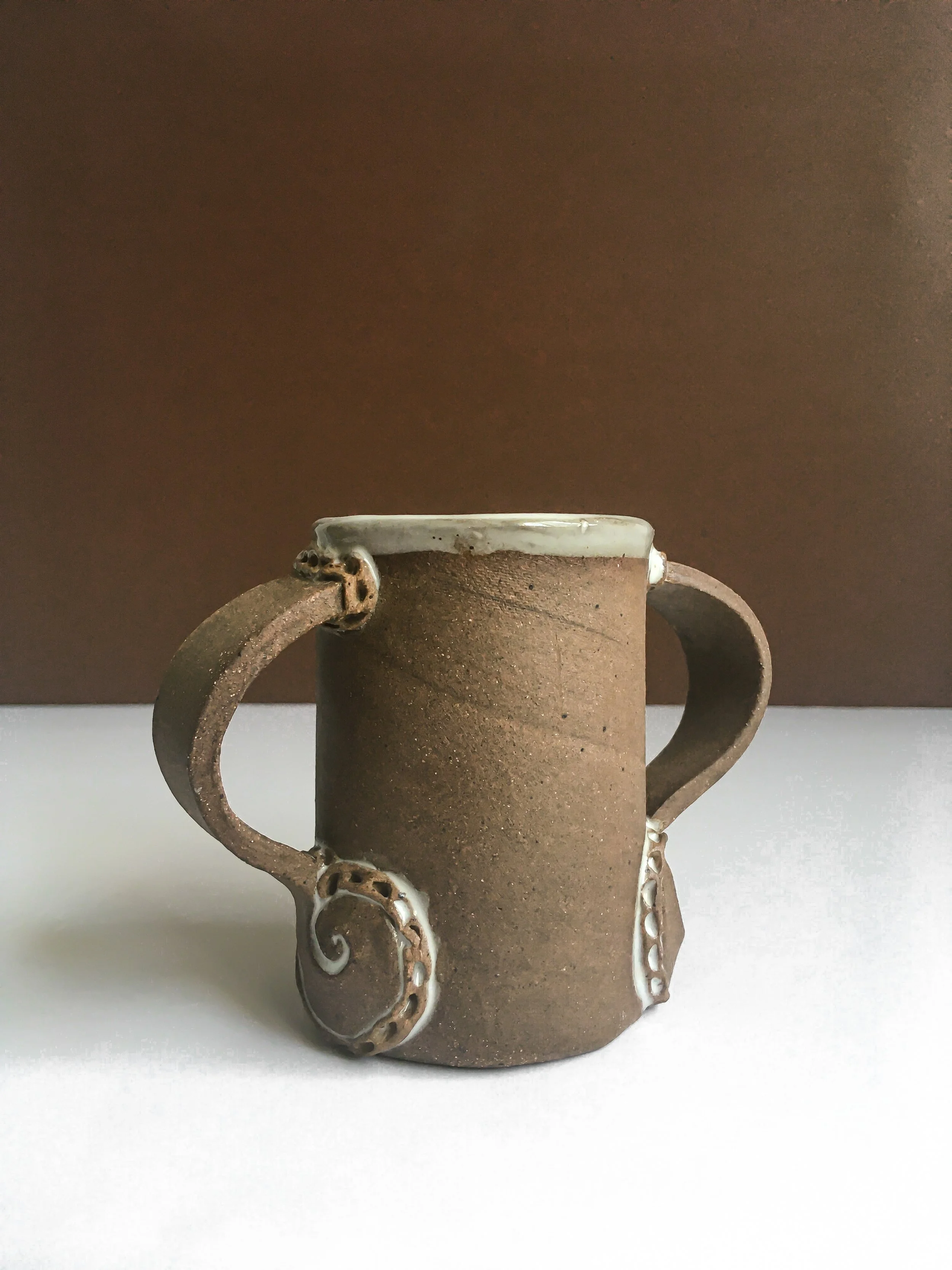 A ceramic mug with two handles, made from a textured, brown clay with decorative white accents near the handles, against a brown and white background.