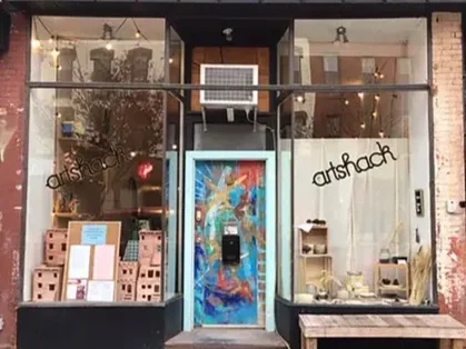 Street view of Artshock storefront with large front windows displaying art supplies and artwork, and a colorful door in the center.