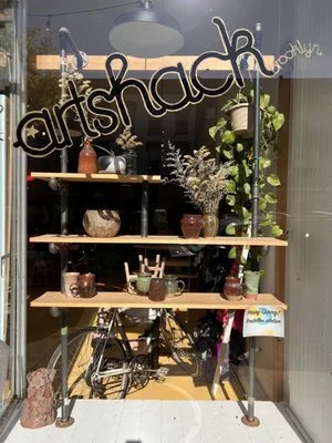 A display window with a wooden table holding various small potted plants, decorative vases, and pottery. Behind the table are shelves with more plants and decorative items. The sign says '@artshackBrooklyn'.
