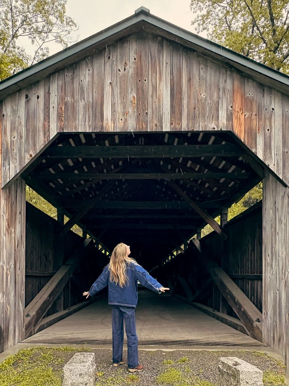 A woman with long blonde hair in a blue jacket and jeans stands with arms outstretched, facing a wooden covered bridge. Soleil Design Co founder and creative director, Sarah Stevens of Massachusetts.