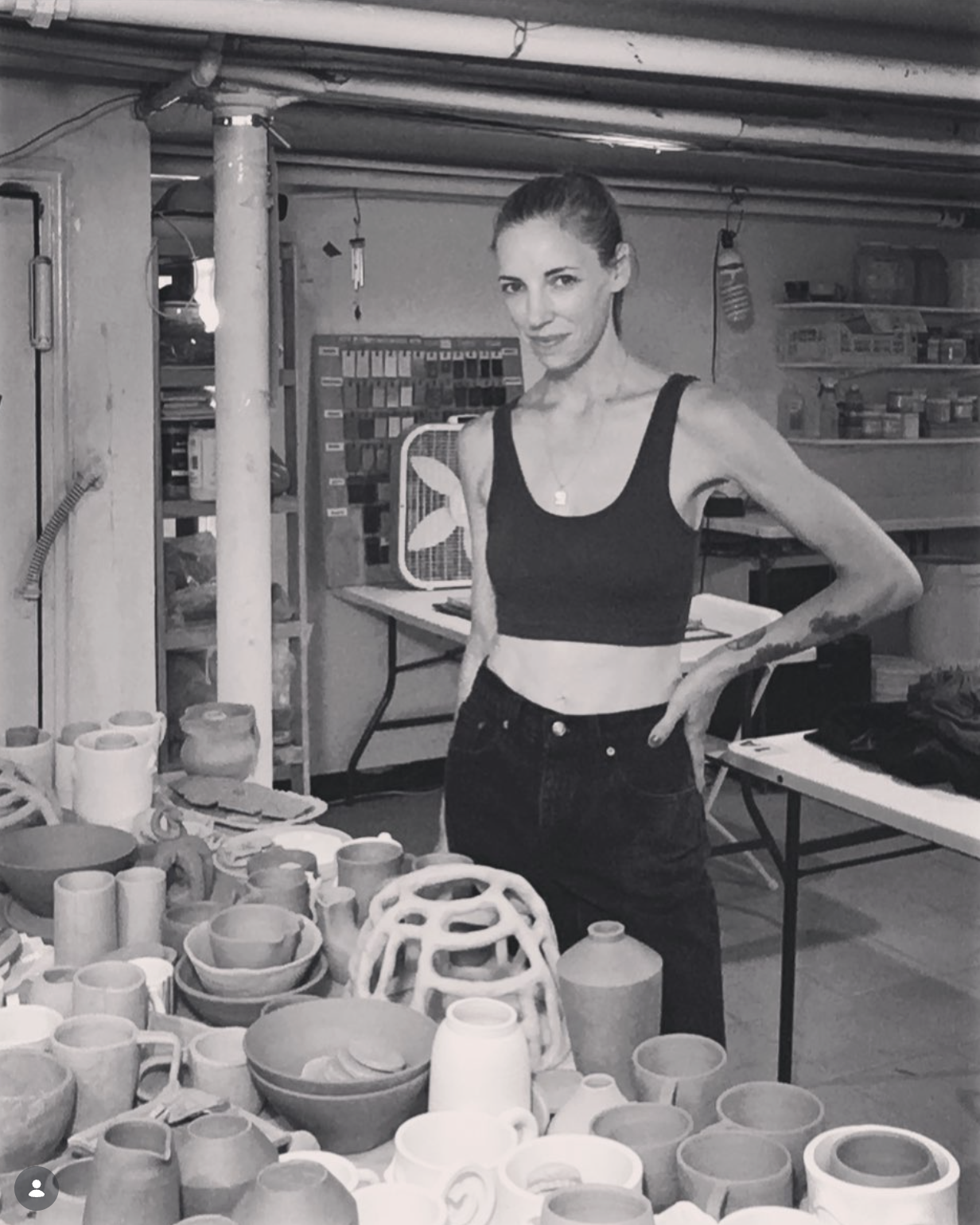 Sarah Stevens with a ponytail and tattoos wearing a black crop top and high-waisted pants, standing at a table filled with various pottery pieces and ceramics in a workshop.
