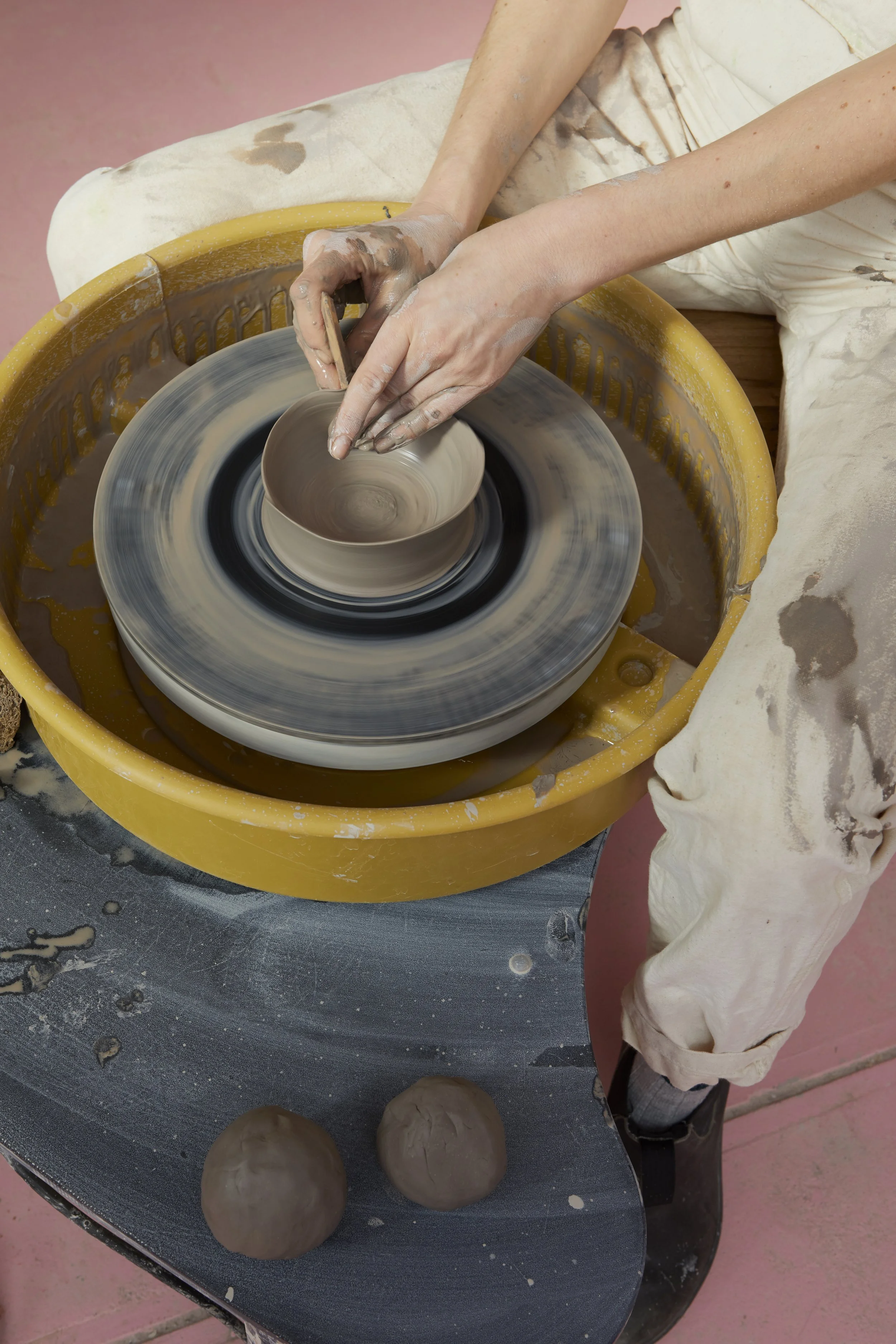 Sarah Stevens shaping clay on a pottery wheel with hands covered in clay, with two clay balls on the table.