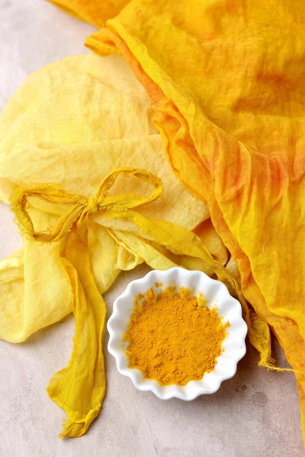 A small white dish filled with bright yellow turmeric powder, placed on a light surface. Around it are yellow fabric cloths tied with a knot, also in shades of yellow.
