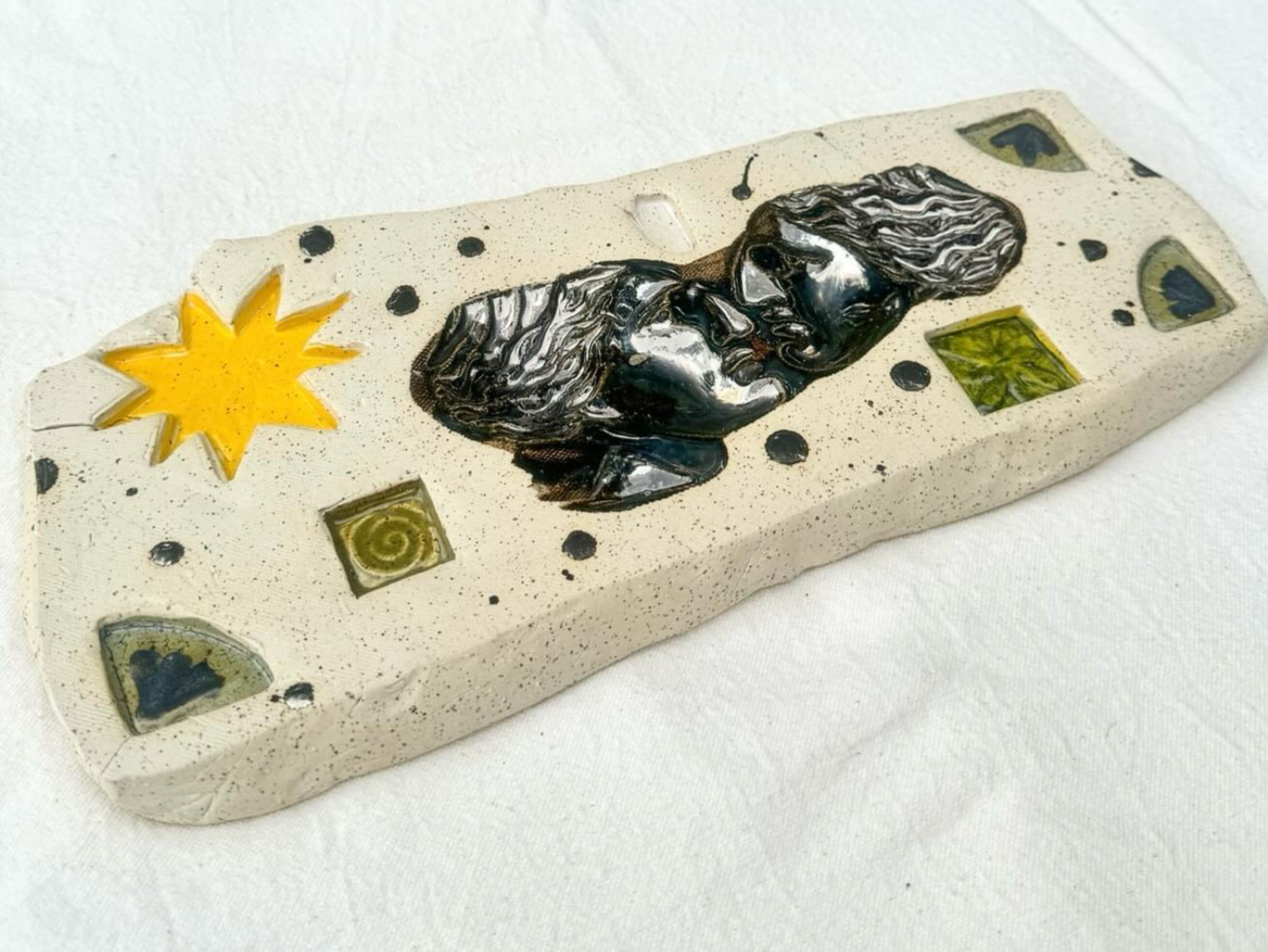 A decorative ceramic tile with abstract and geometric designs, including a yellow star or sun shape, black dots, and green square and triangle accents, in a mix of black, yellow, and green colors.