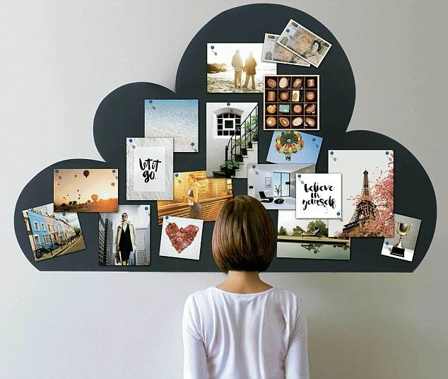 A woman looking at a cloud-shaped collage of photos, notes, and tickets on a wall.