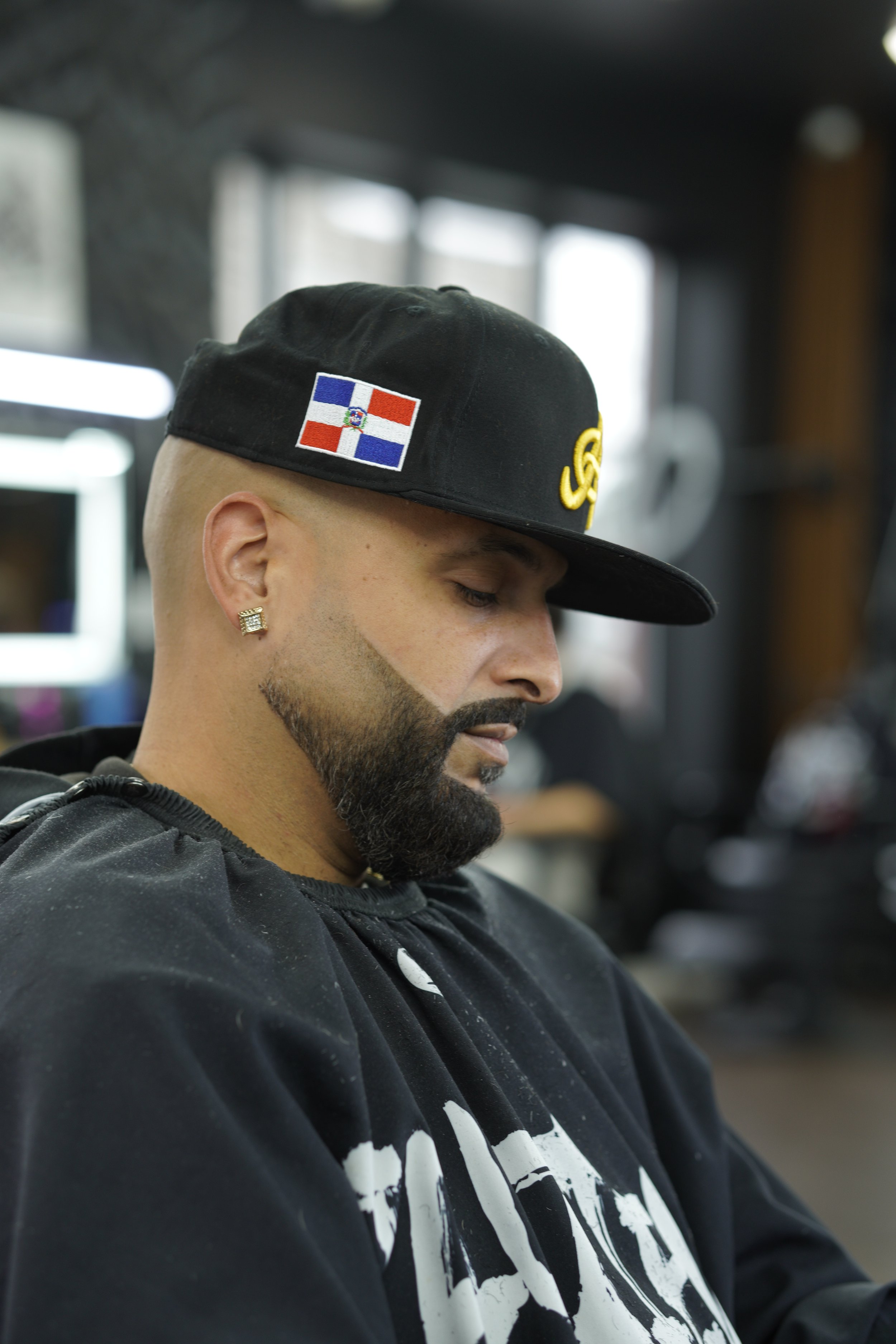 A man with a beard and gold earring wearing a black baseball cap with a Dominican Republic flag patch, sitting indoors with a black t-shirt with white graphics, eyes closed or looking downward.