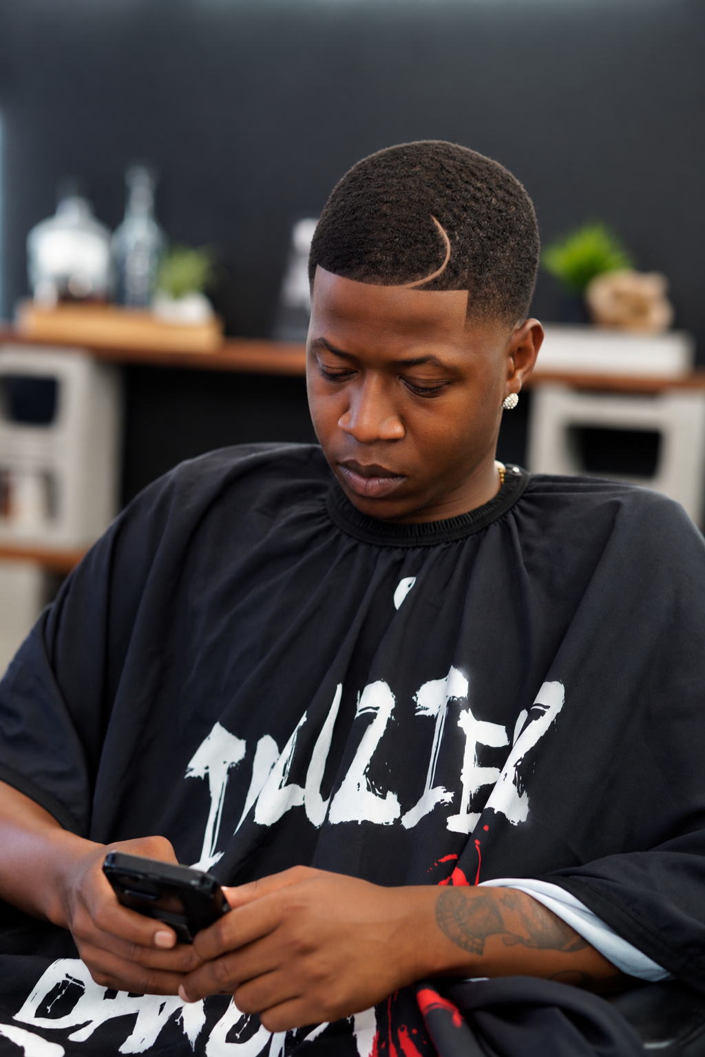 A young man with a fade haircut looking at his phone in a barbershop, wearing a black barber cape with white and red text.