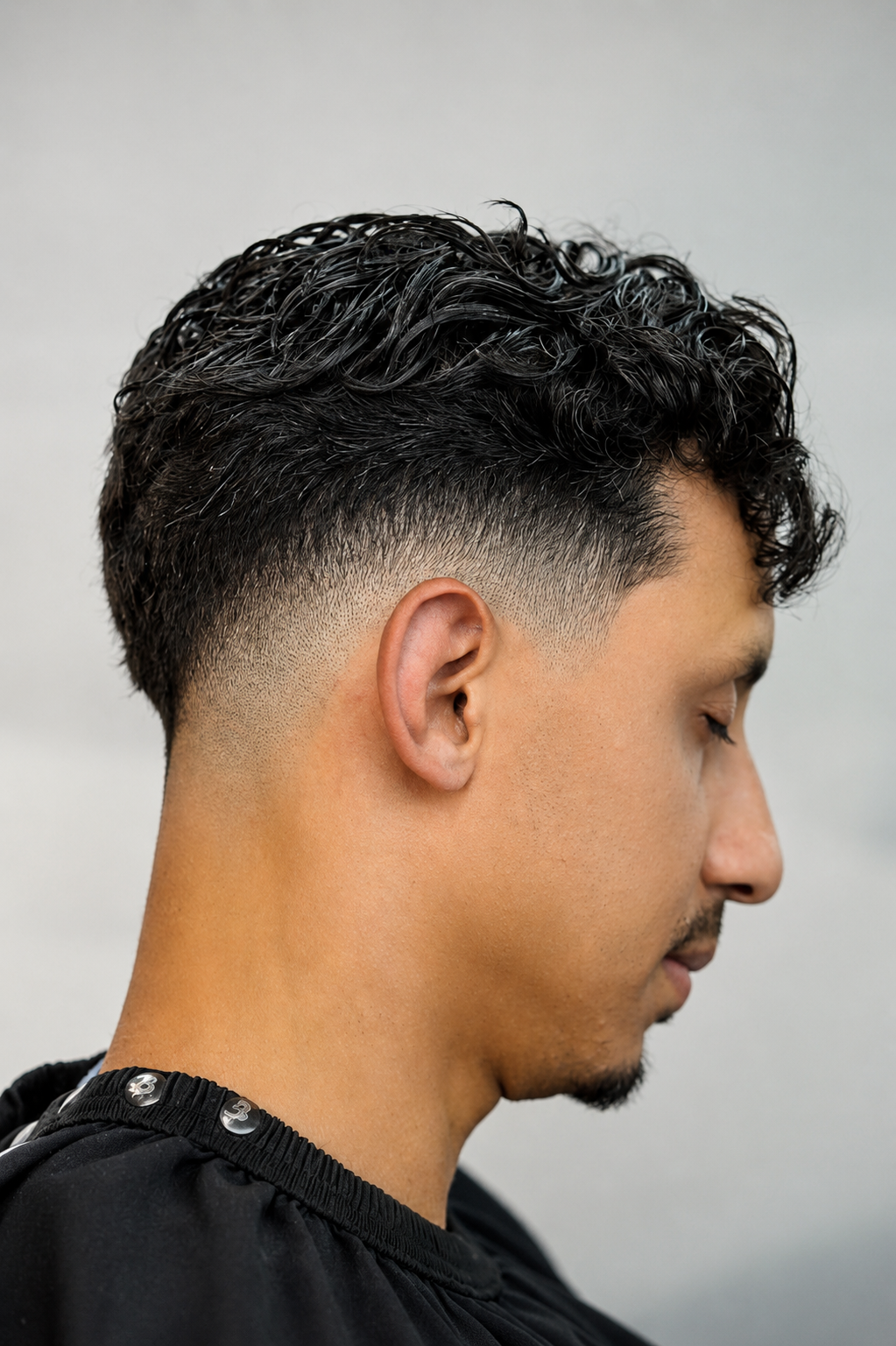 Side profile of a man with curly dark hair and a short fade haircut, wearing a black shirt.