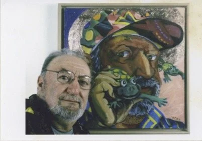 A man with glasses and a beard takes a selfie in front of a colorful self portrait of him wearing a hat with a frog in his hand.