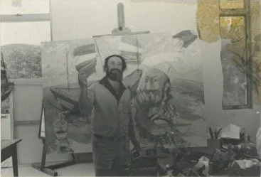 An artist standing in front of a large landscape painting in an art studio.