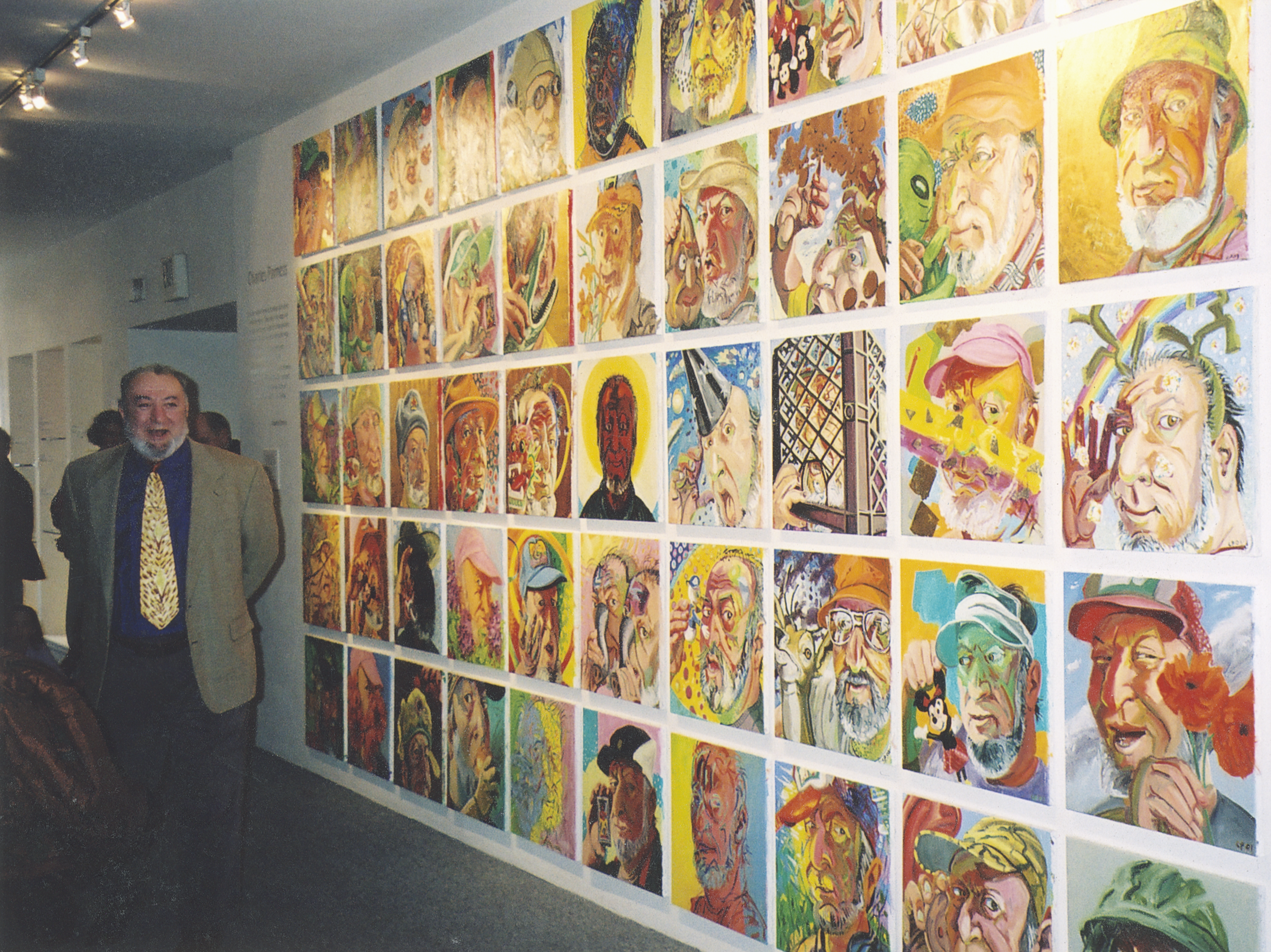 A man in a beige blazer and patterned tie stands in front of a wall display of colorful portrait paintings at an art exhibition.