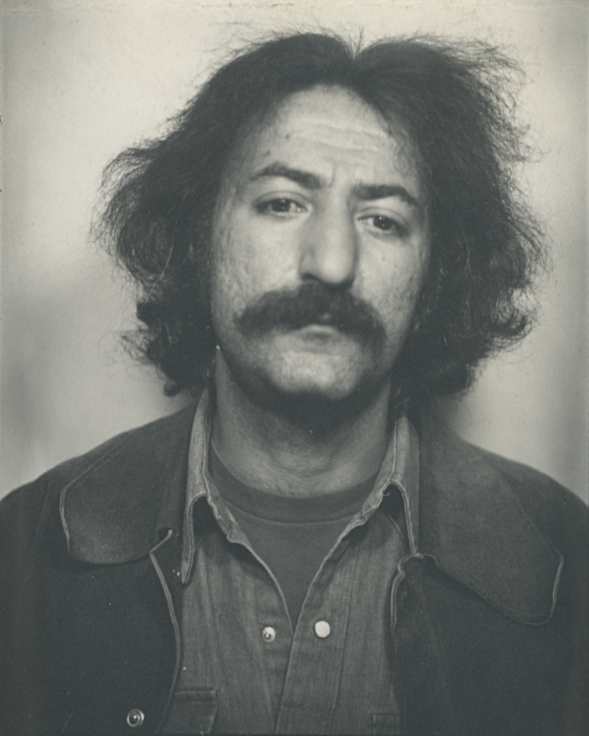 Black and white portrait of a man with thick, curly hair and a mustache, wearing a denim shirt and a jacket.