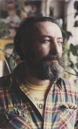 A man with a beard and long dark hair, wearing a plaid shirt, looking to the side indoors with plants in the background.