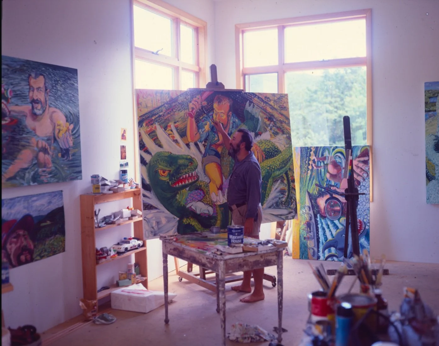 An artist is painting a large colorful canvas in a bright studio with several other paintings on the walls and a shelf with model cars, paint cans, and art supplies.