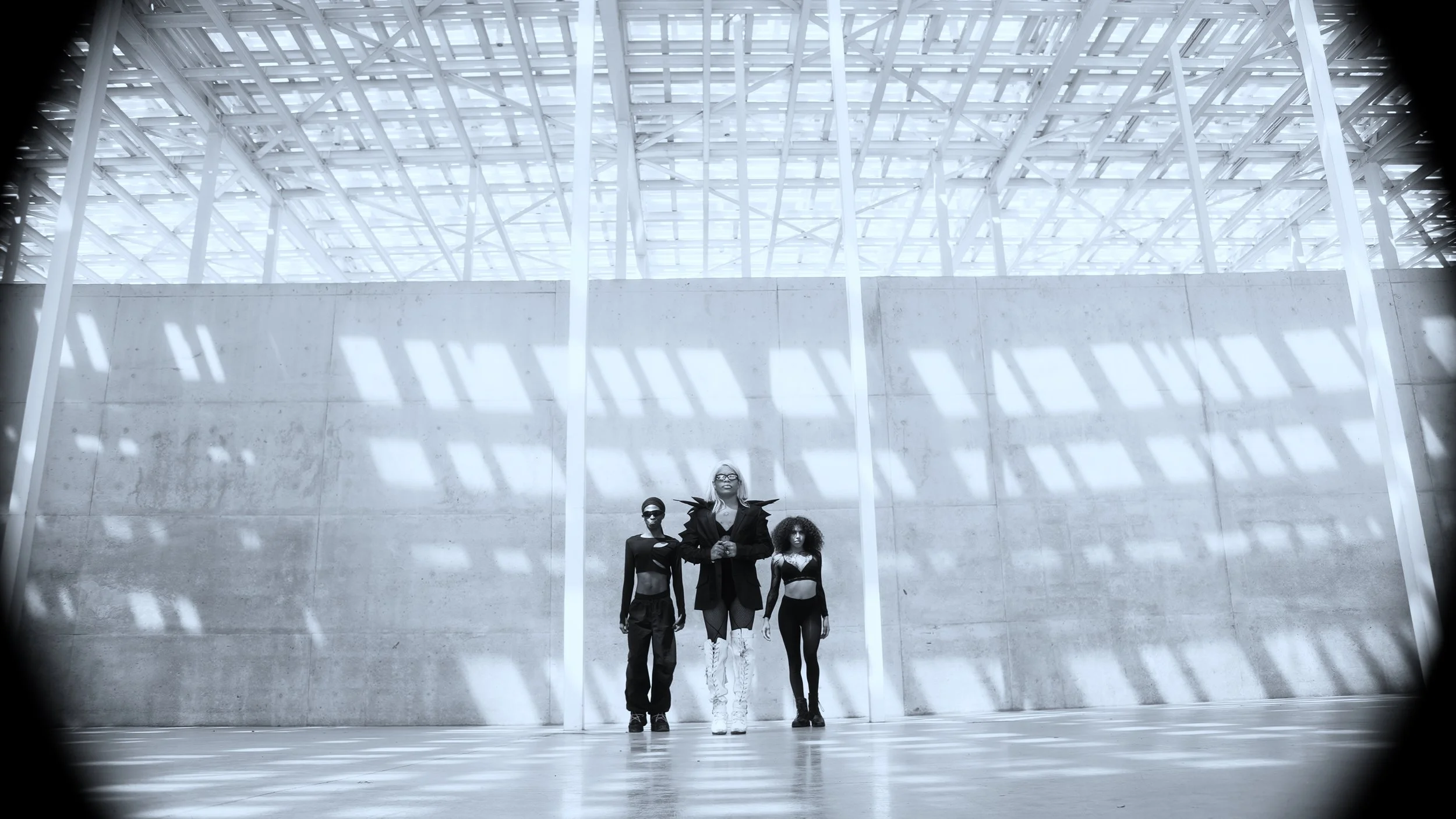 Three fashionably dressed individuals standing in a sunlit, industrial space with concrete walls and grid-patterned ceiling.