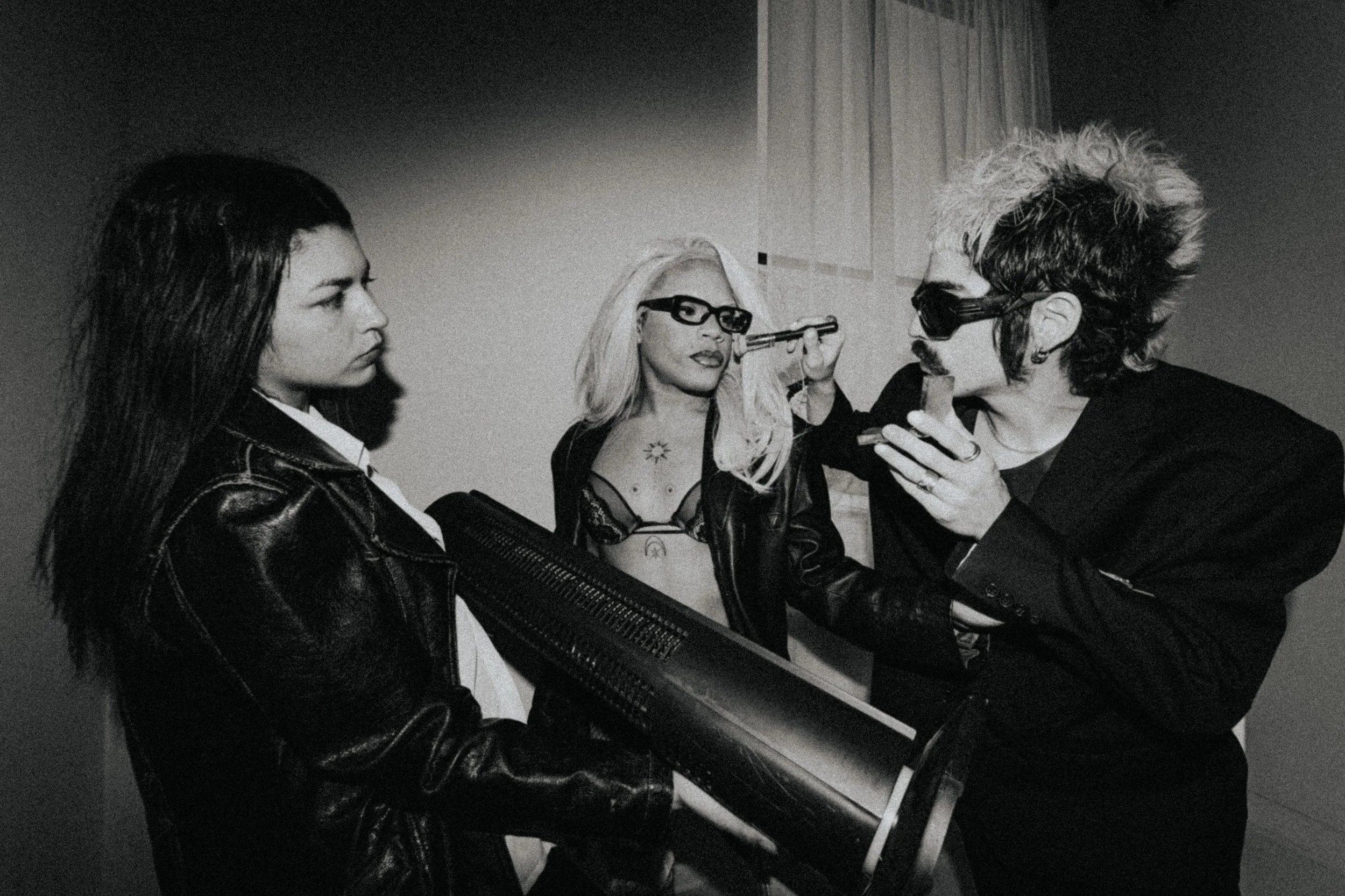 Black and white photo of three people wearing dark sunglasses and leather jackets, with one person holding a fan. The central person has blonde hair and is wearing a revealing top. They appear to be having an animated conversation.