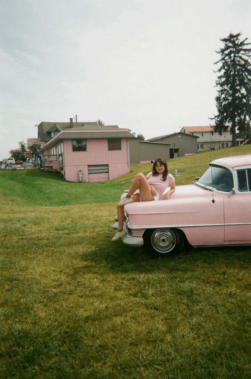 Person sitting on the hood of a vintage pink car, near a pink building in a grassy area.