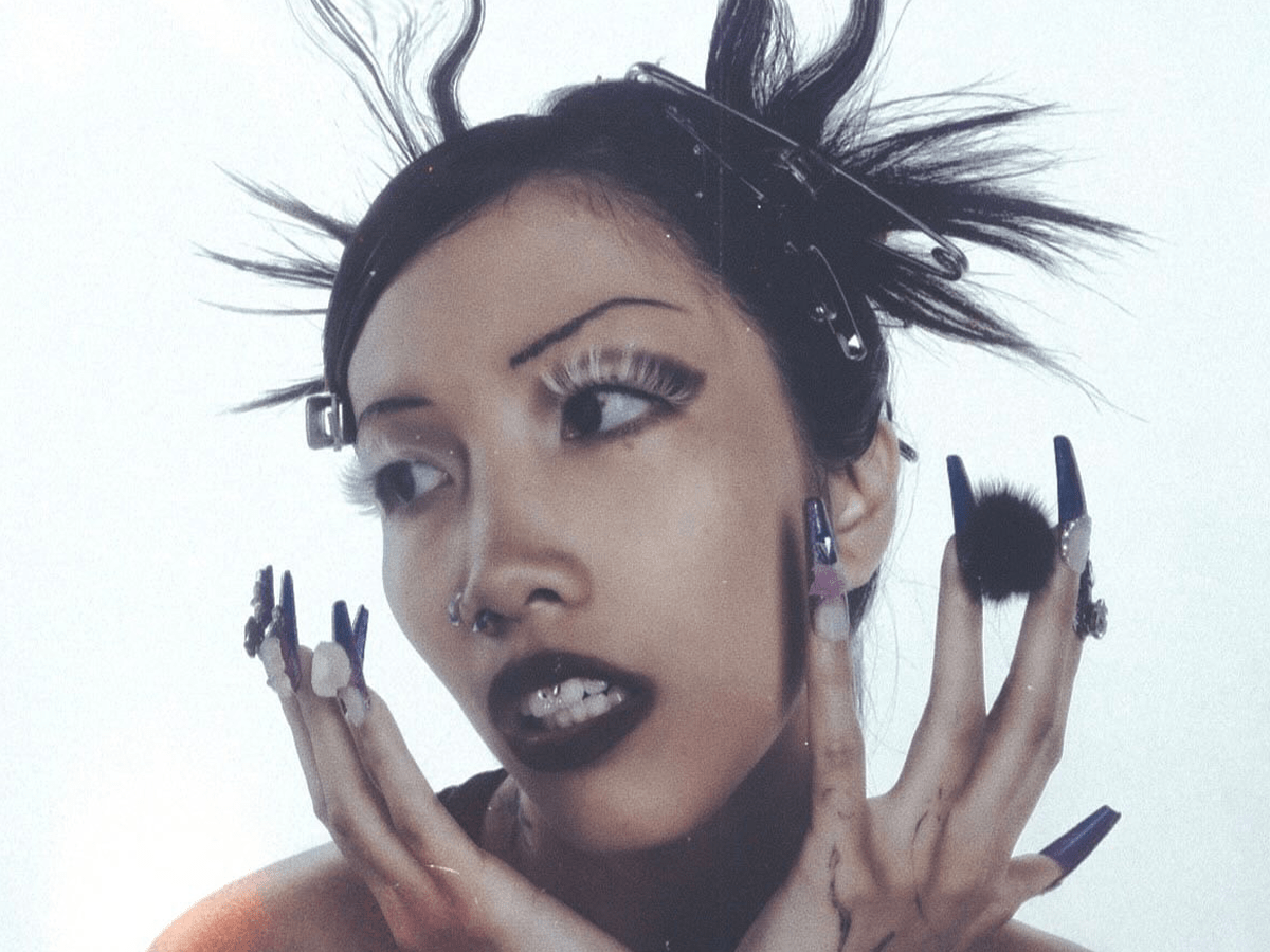 Person with spiked hair and elaborate nail art, wearing heavy makeup and facial piercings, looking to the side.