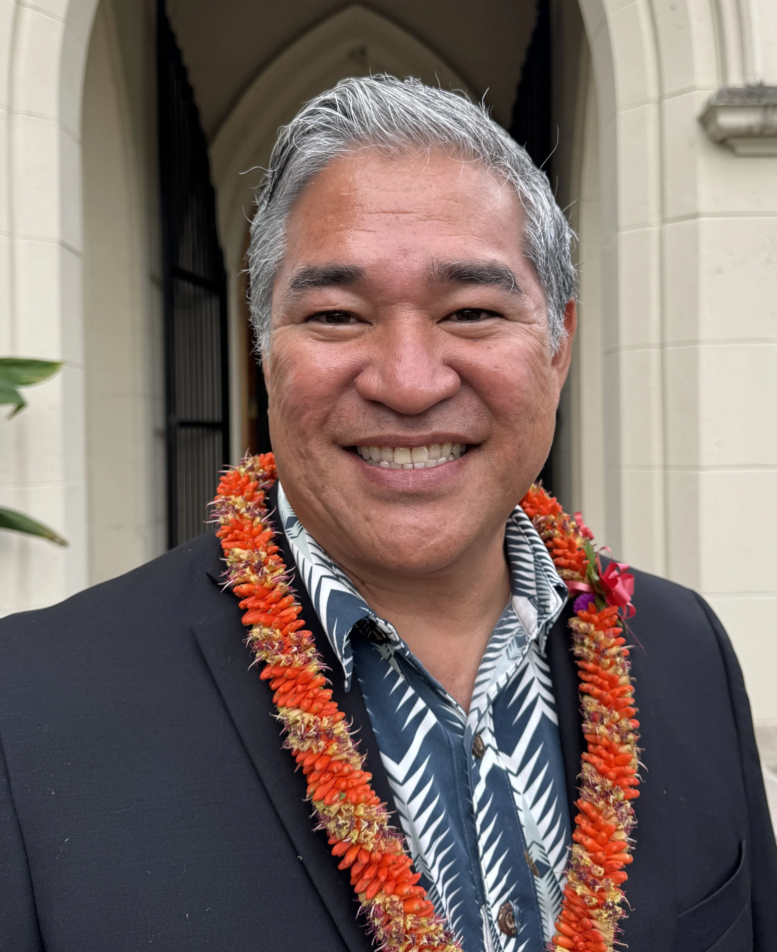 Trustee, Daniel M. Goya smiling in front of the Lunalilo Mausoleum, wearing a lei and formal attire