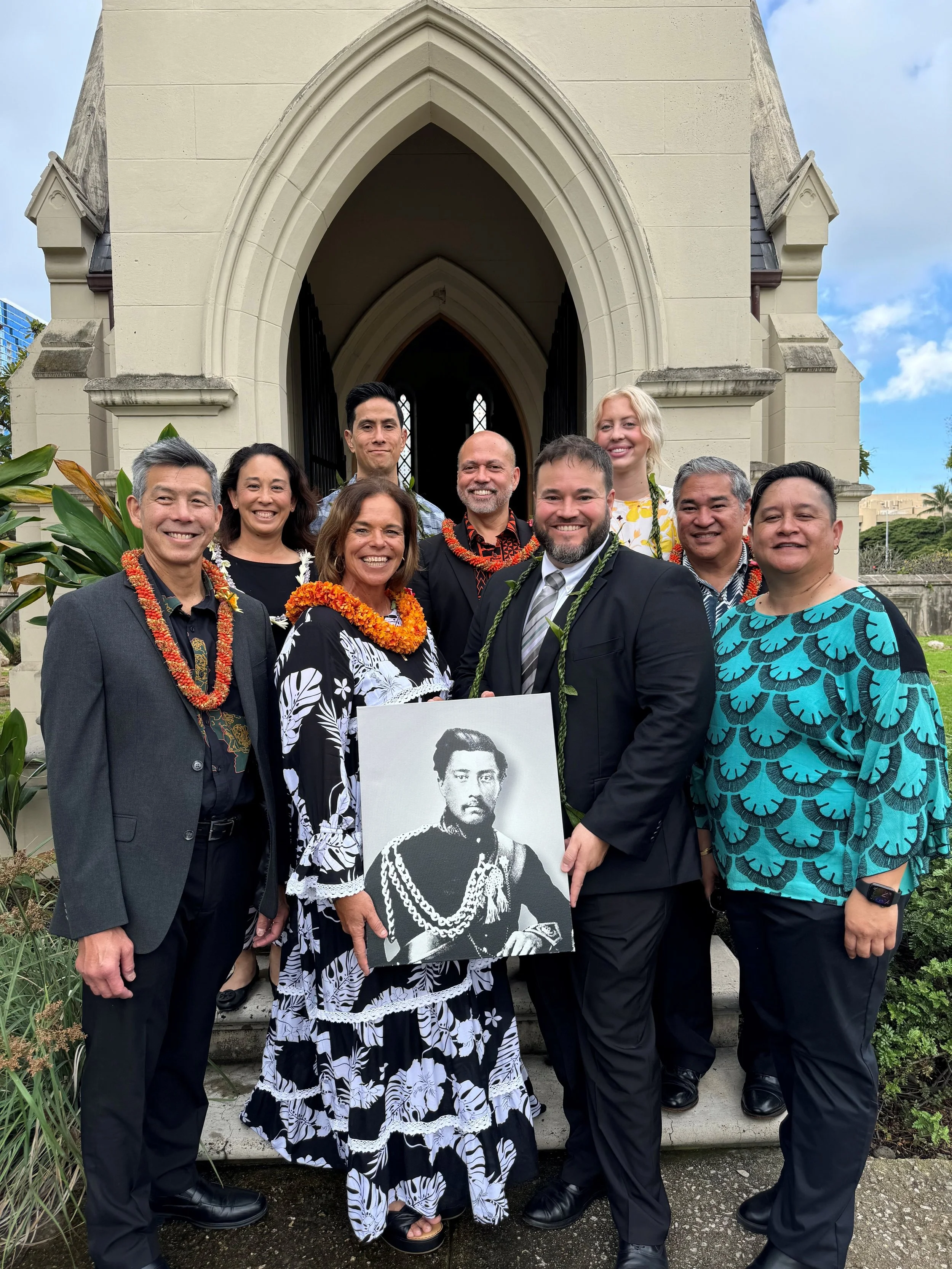 Smiling trustees and leadership members of the King Lunalilo Trust posing in front of the Lunalilo Mausoleum holding a portrait of King Lunalilo