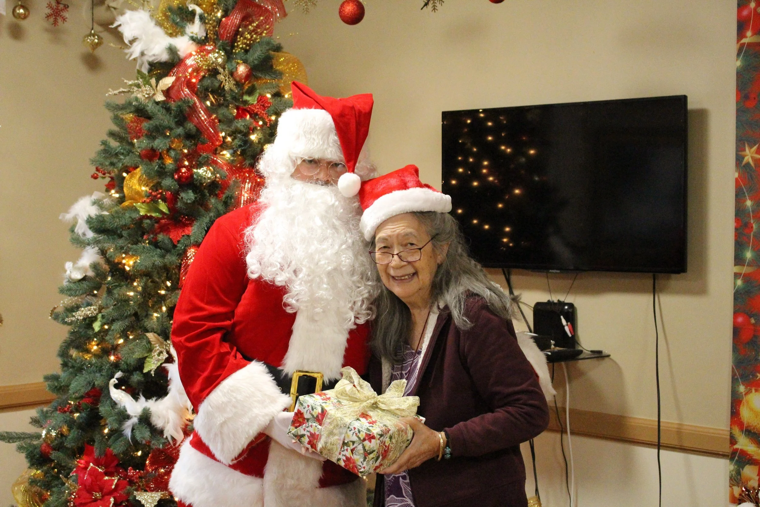 Santa and a kūpuna (elderly resident) posing with a present next to a Christmas tree during a holiday celebration at Lunalilo Home.