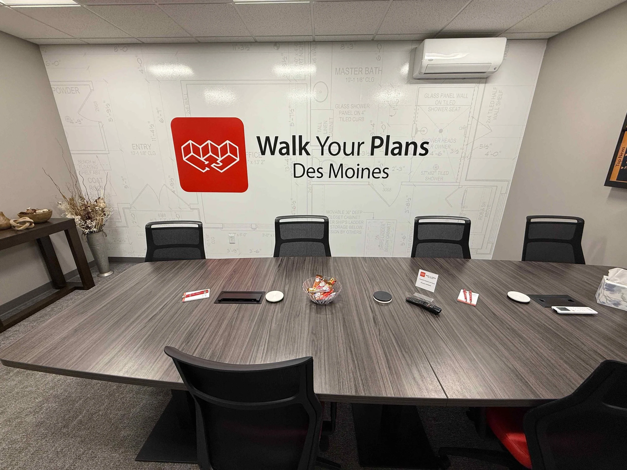 A conference room with a large oval table surrounded by black chairs, a wall with the logo of Walk Your Plans Des Moines and architectural blueprints, and various items on the table including pens, a remote control, a small bowl of candy, and a tissue box.