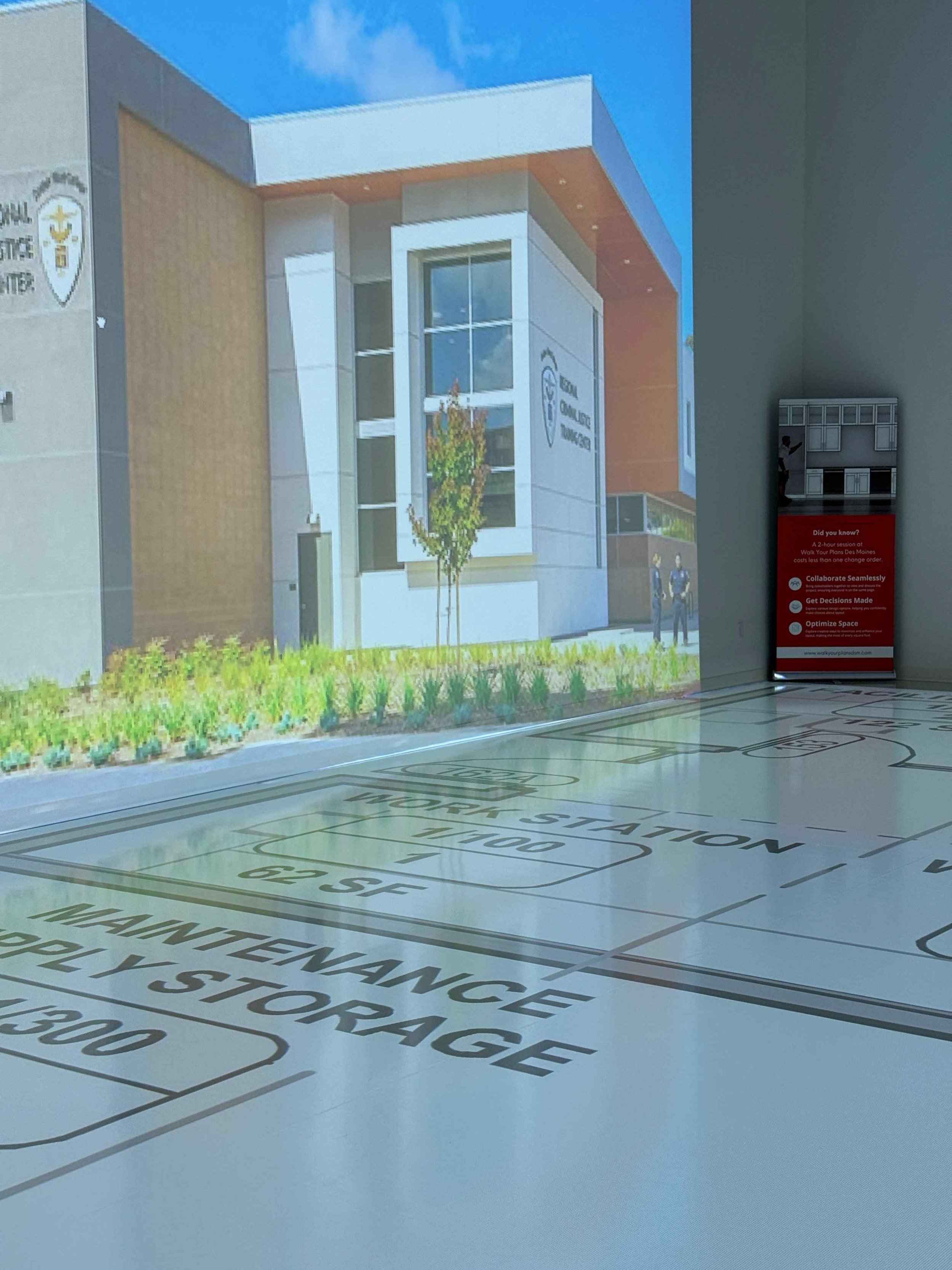 A digital rendering of a modern building with the logo and name of a school, wall and window details, with a landscaped area in front. The image is projected onto a floor with floor plans, emergency routes, and safety instructions.