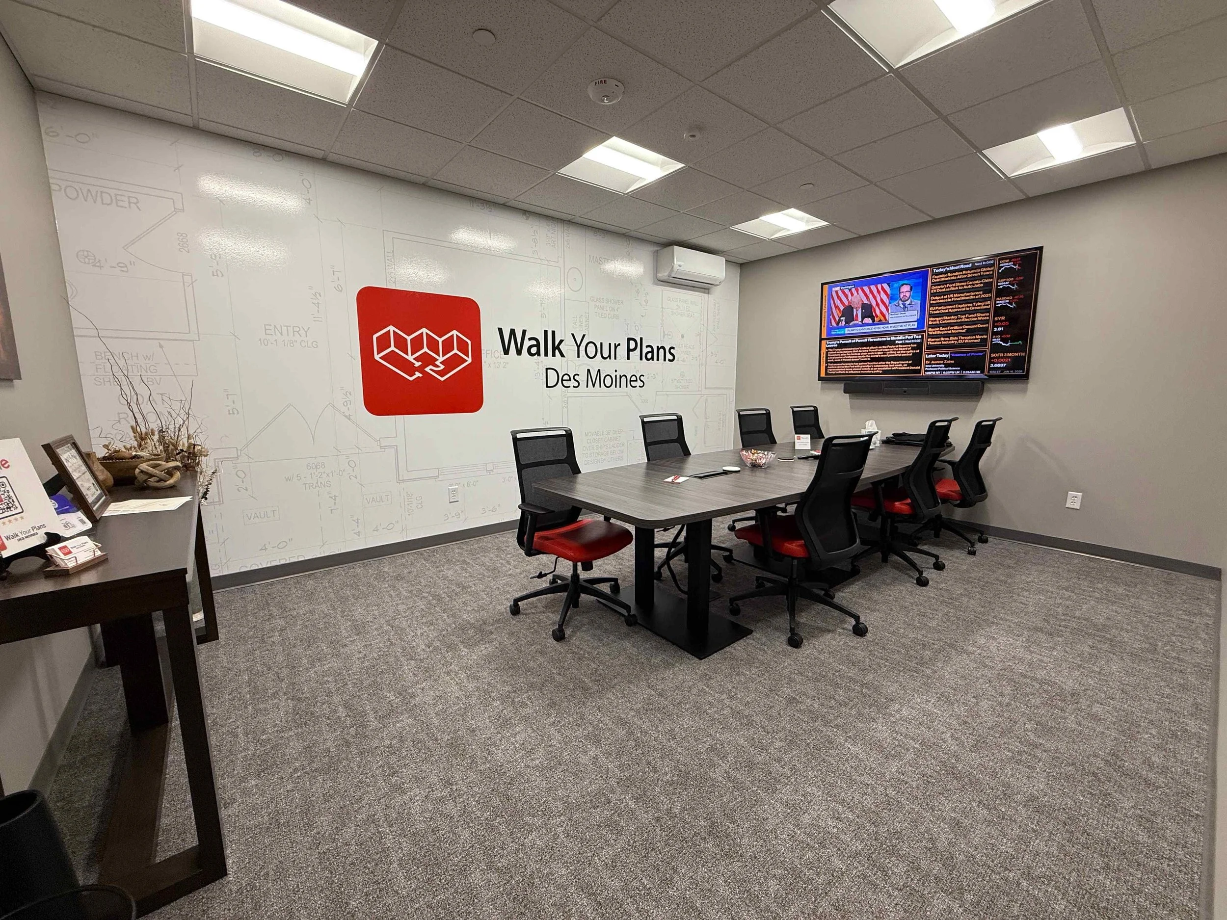 A conference room with a large table surrounded by six black and red office chairs, a wall with a logo and the text 'Walk Your Plans Des Moines', a wall-mounted TV displaying news, and a small table with brochures and decorative items.