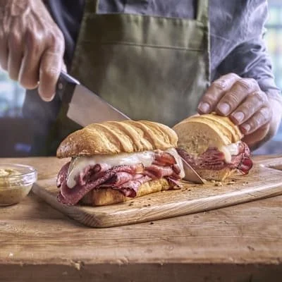 Person slicing a sandwich with roast beef and melted cheese on a wooden cutting board.