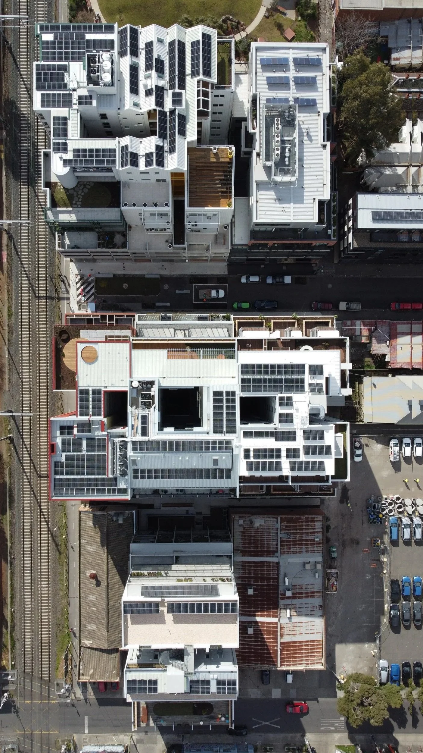 Aerial view of modern buildings with solar panels on rooftops, adjacent to railway tracks and a parking lot.