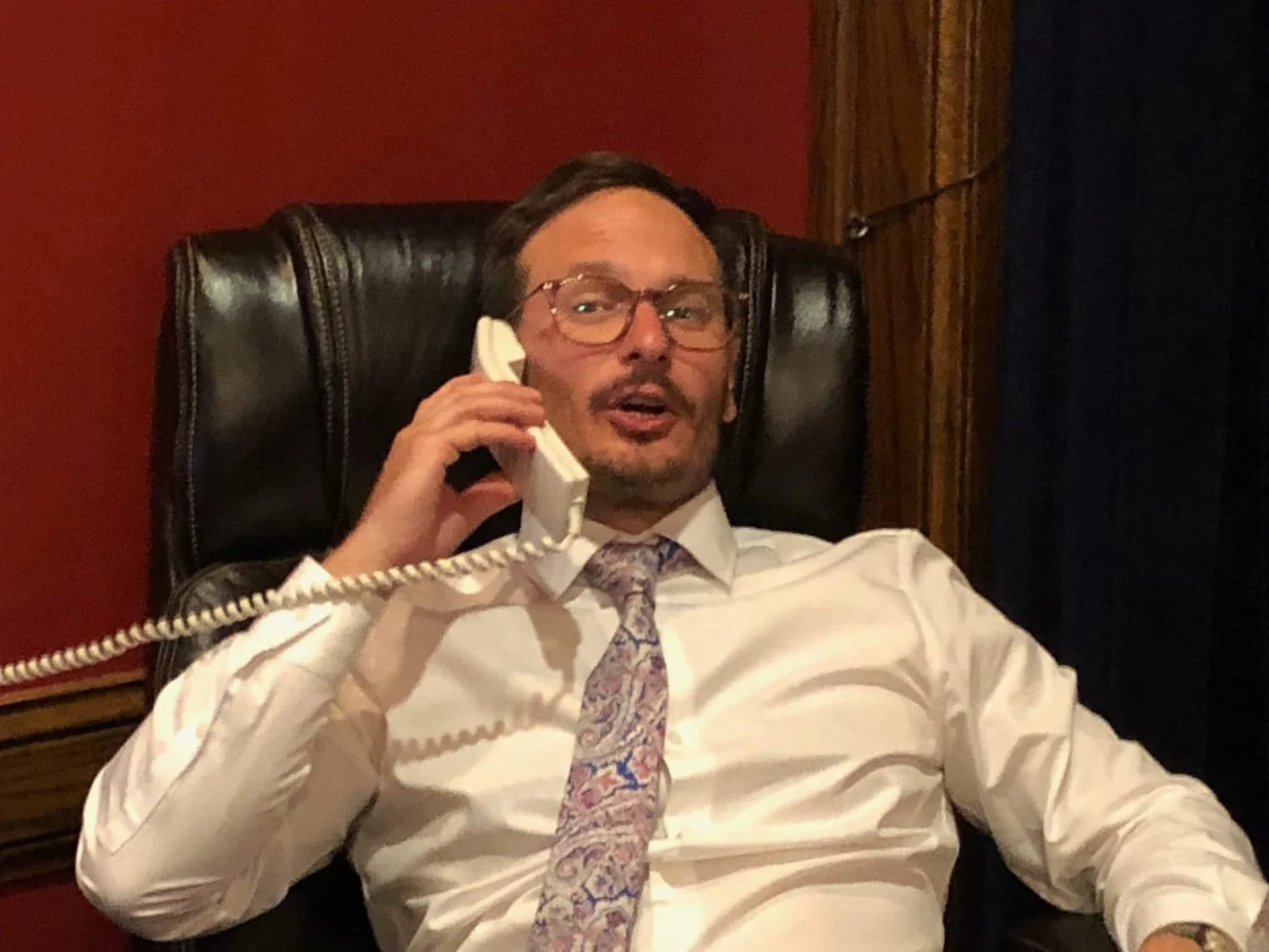 A man wearing glasses and a white shirt with a colorful tie, sitting in a leather chair, talking on a corded phone.