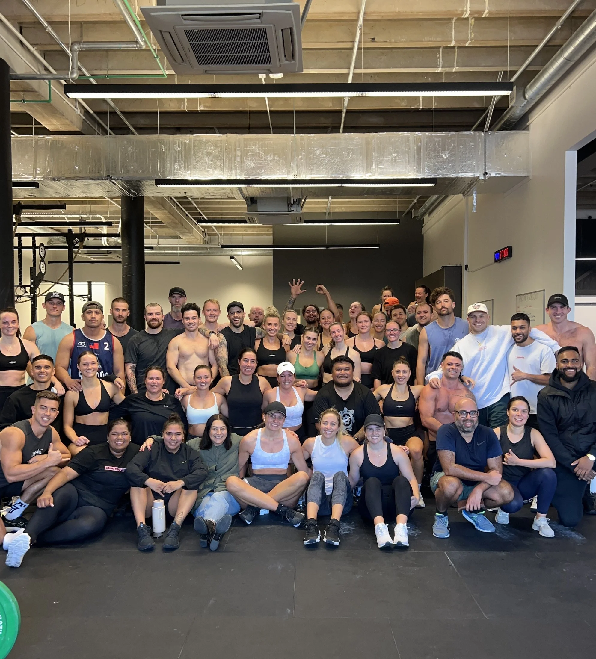 A large group of people posing for a photo in a gym setting, wearing athletic clothing, some shirtless, against a background of fitness equipment.