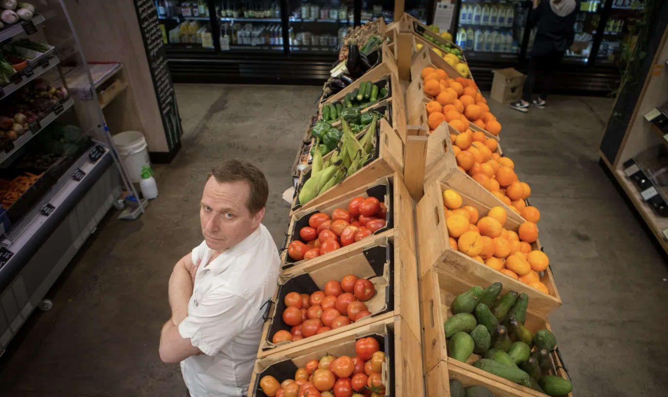 A man in an organic grocery store aisle with arms crossed, looking up at the camera. He is next to wooden bins filled with tomatoes, cucumbers, oranges, and avocados.