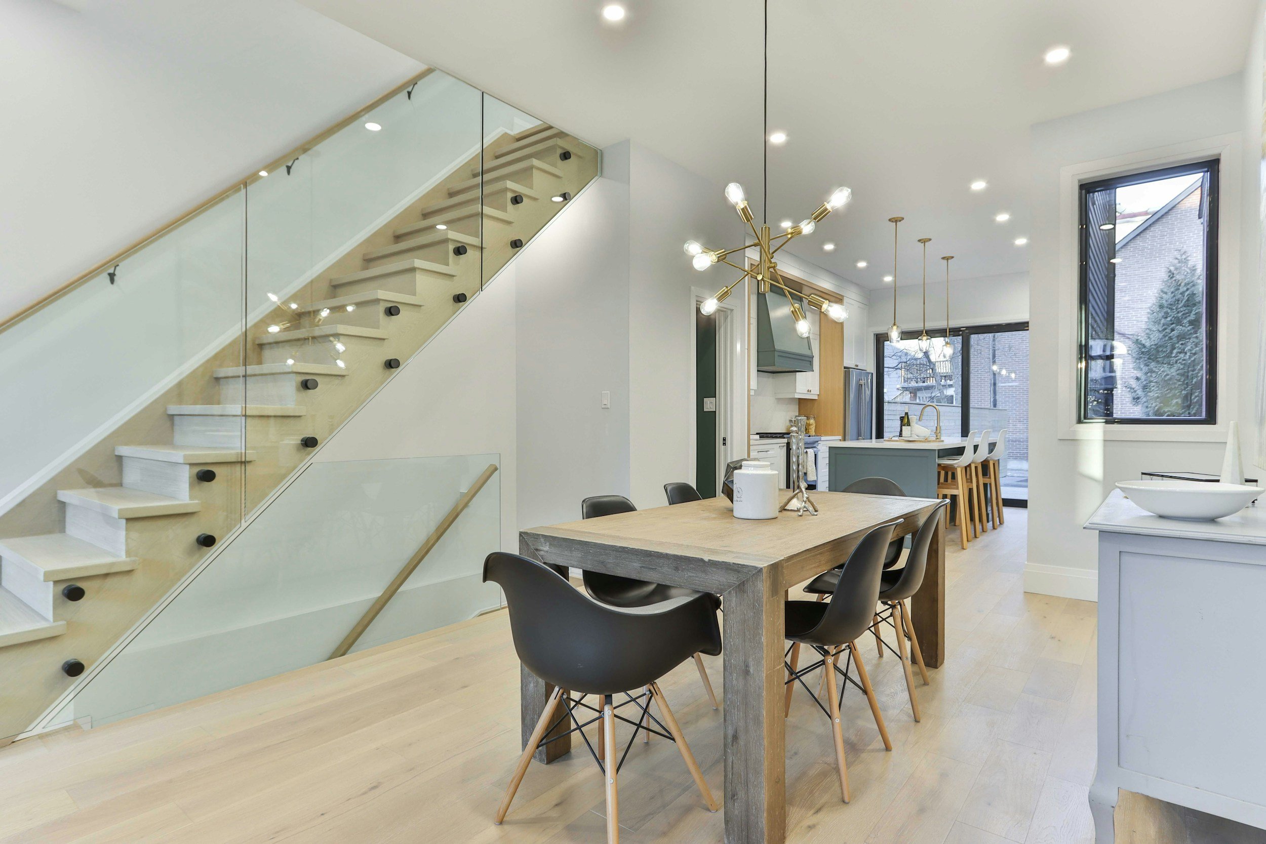 Modern open-concept kitchen and dining area with light wood flooring, a wooden dining table surrounded by black chairs, a contemporary brass chandelier, and large windows letting in natural light.