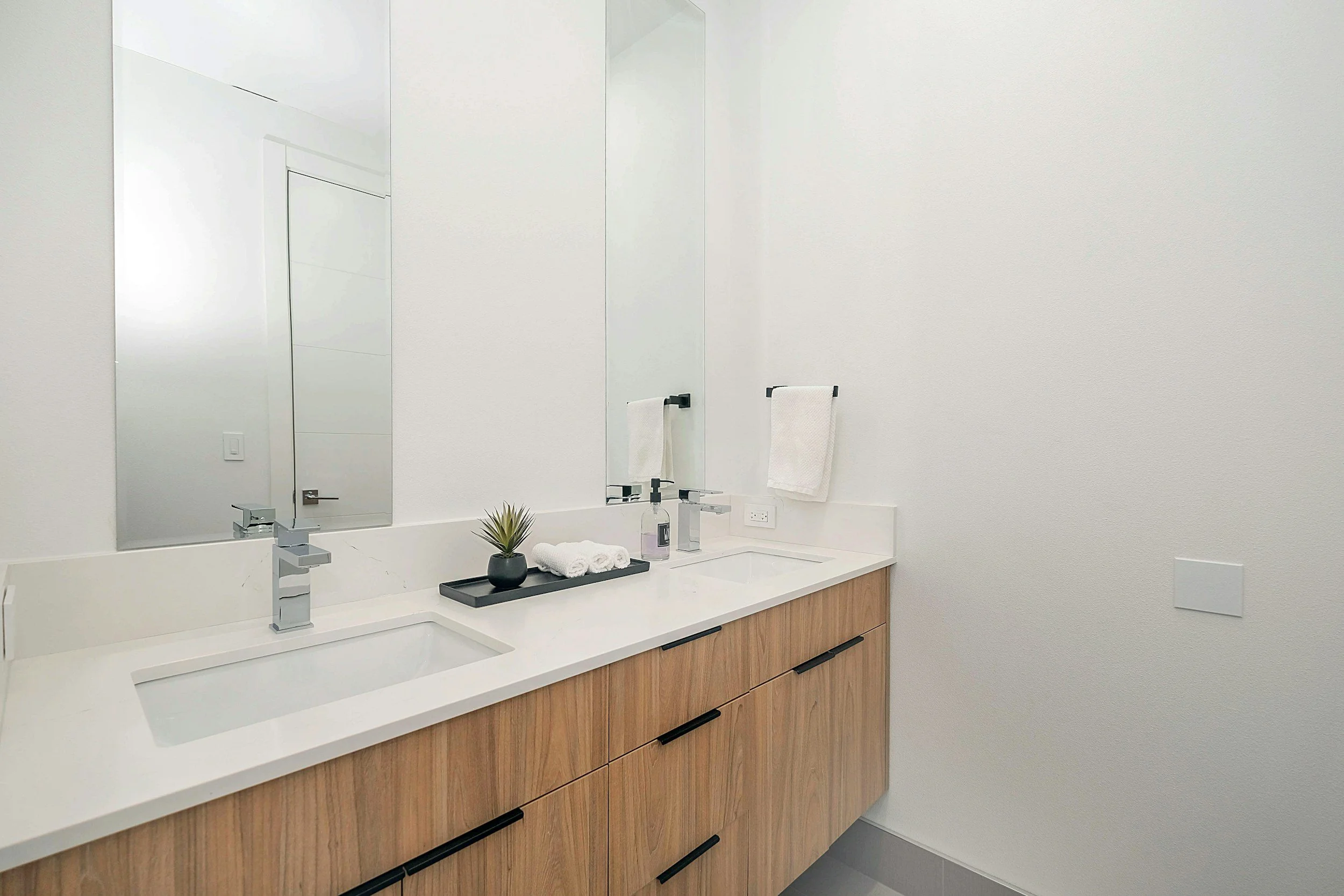 Modern bathroom with dual sinks, large mirror, white countertop, wooden cabinet, towel rack with white towels, small potted plant, soap dispenser, and minimalist decor