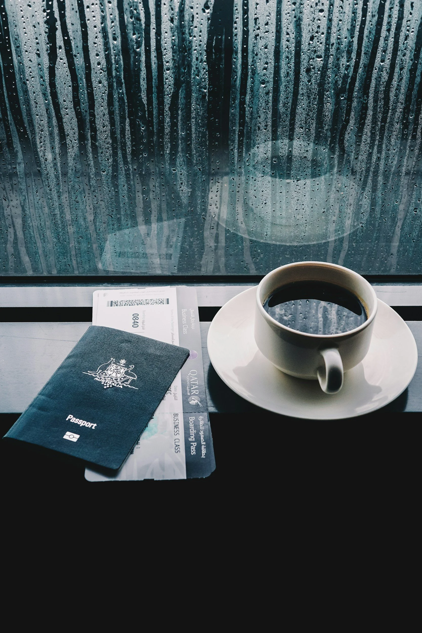 A coffee cup on a saucer, a passport, and boarding passes on a window sill with raindrops on the window.