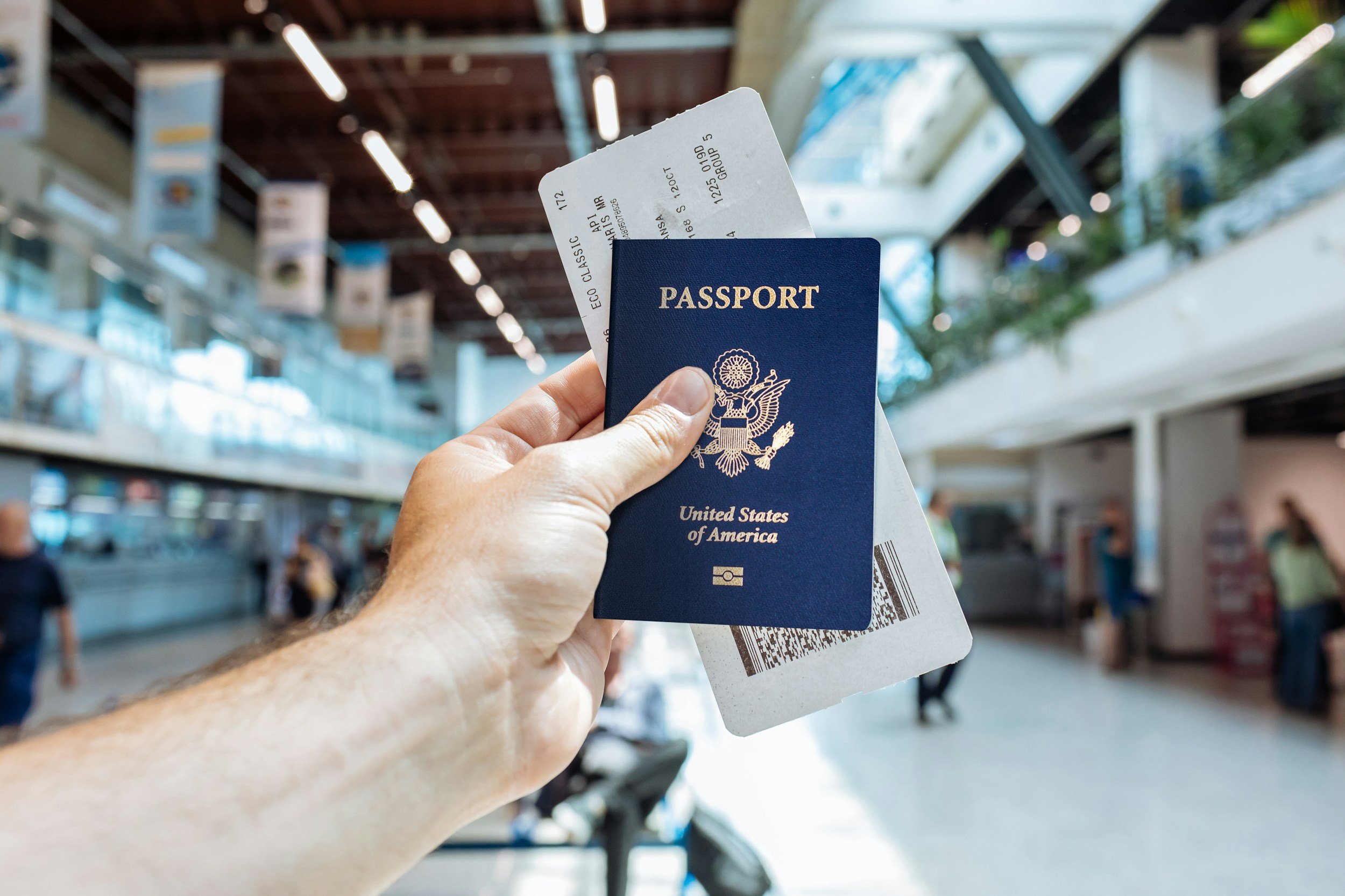 A hand holding a United States passport, a boarding pass, and a flight ticket inside an airport terminal with people walking and baggage areas in the background.