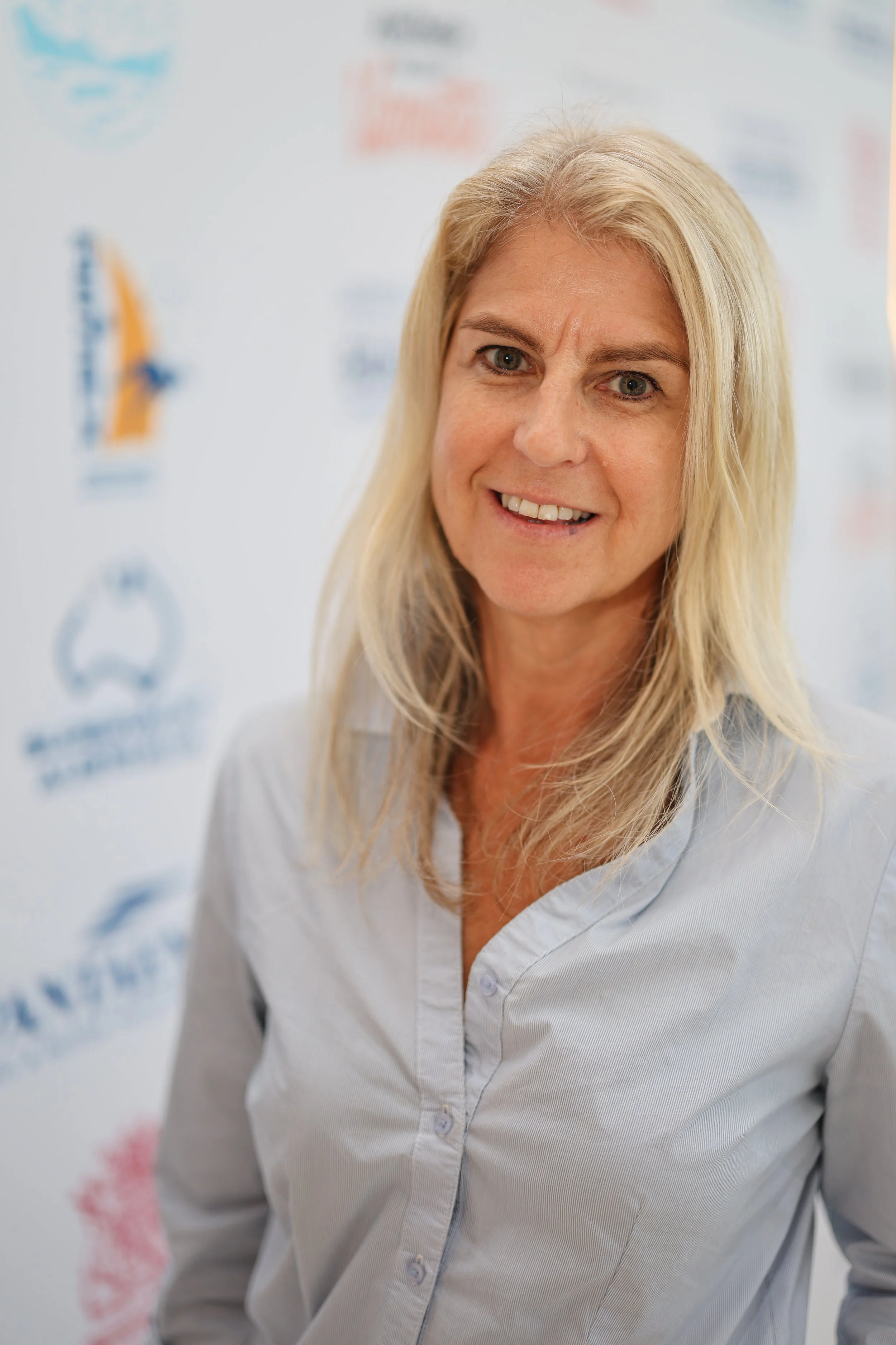 A woman with blonde hair smiling, wearing a light-colored shirt, standing in front of a backdrop with logos and text.
