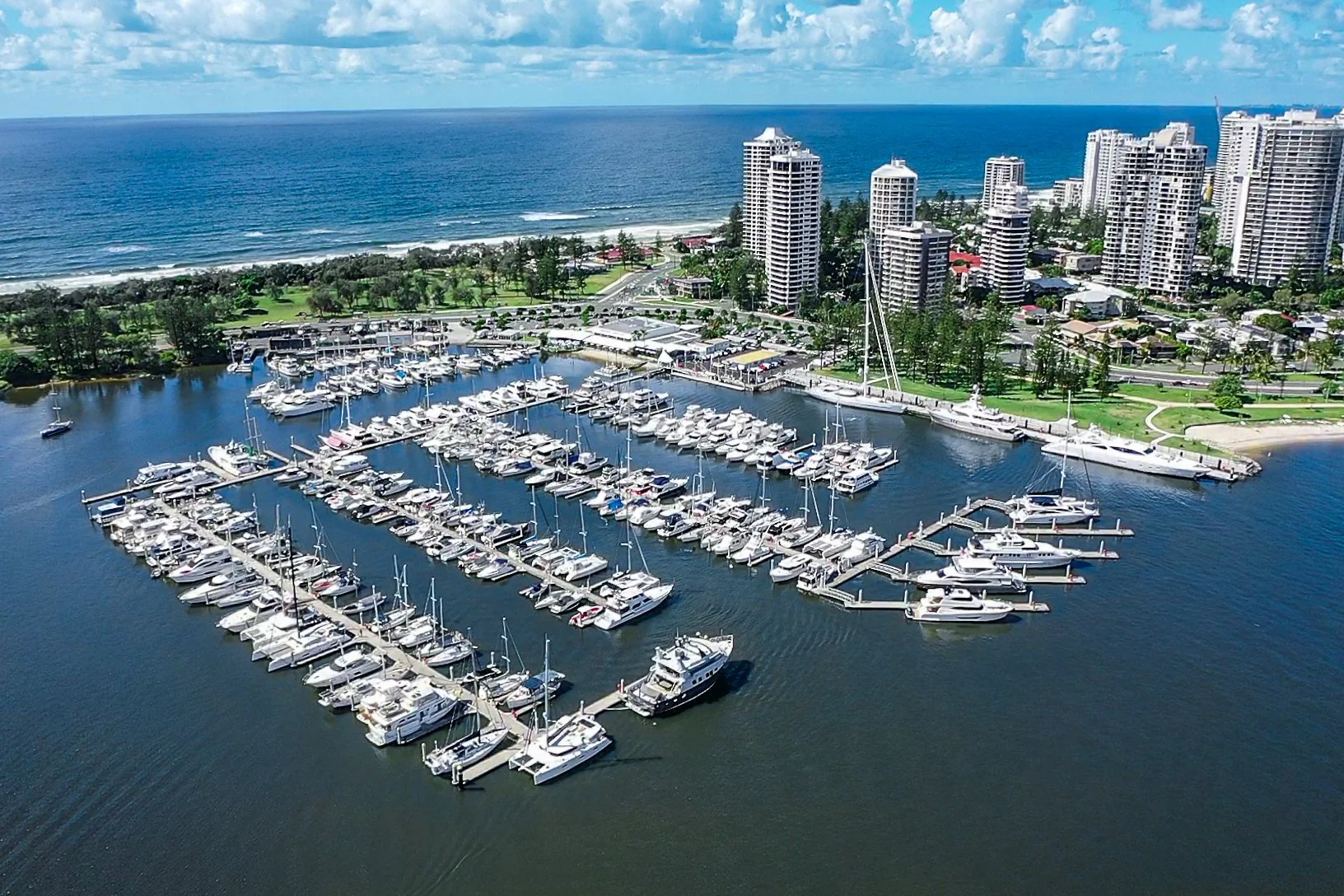 Aerial view of a marina with many boats docked, a city skyline with high-rise buildings, a beach, and the ocean under a partly cloudy sky.
