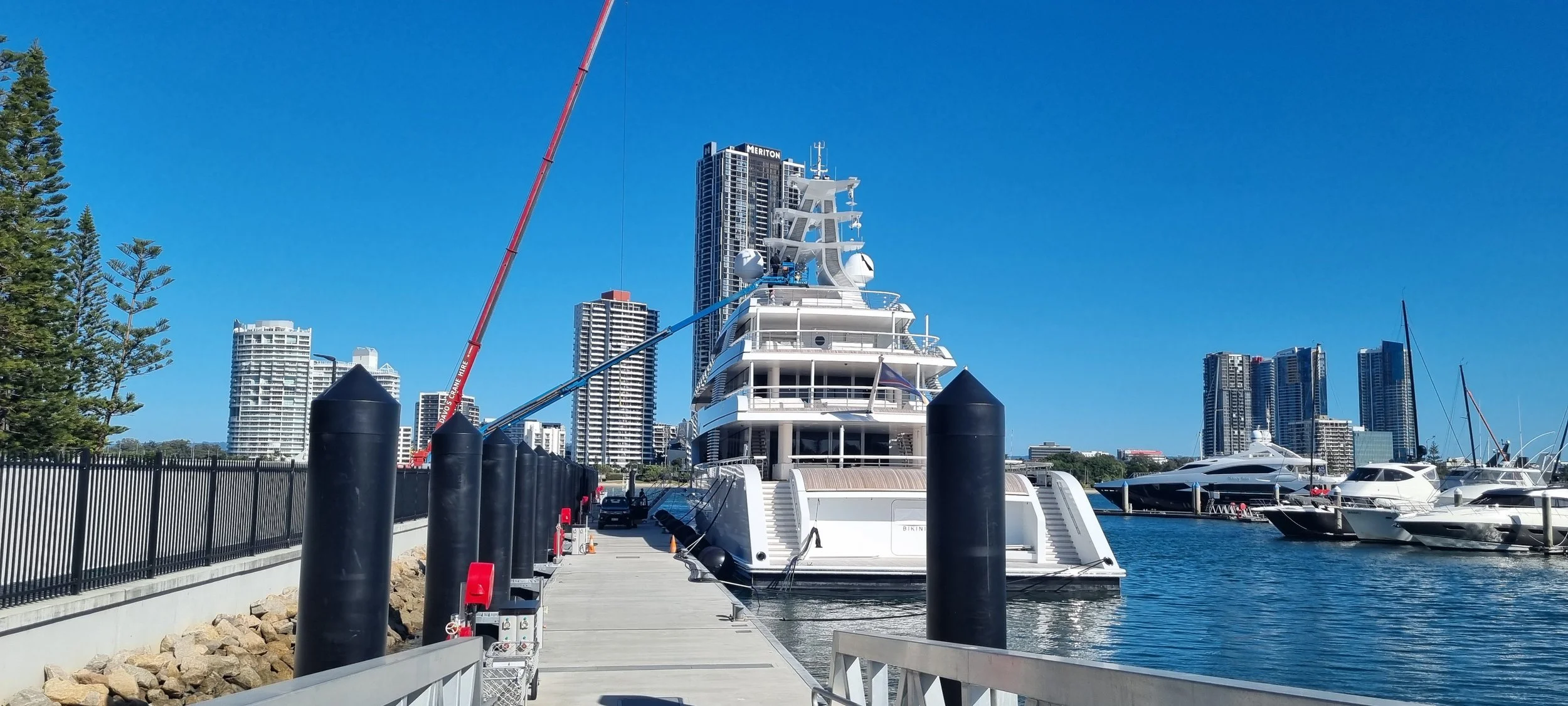 A marina with large yachts docked, a floating concrete dock with black bollards, and high-rise buildings in the background on a clear blue sky day.