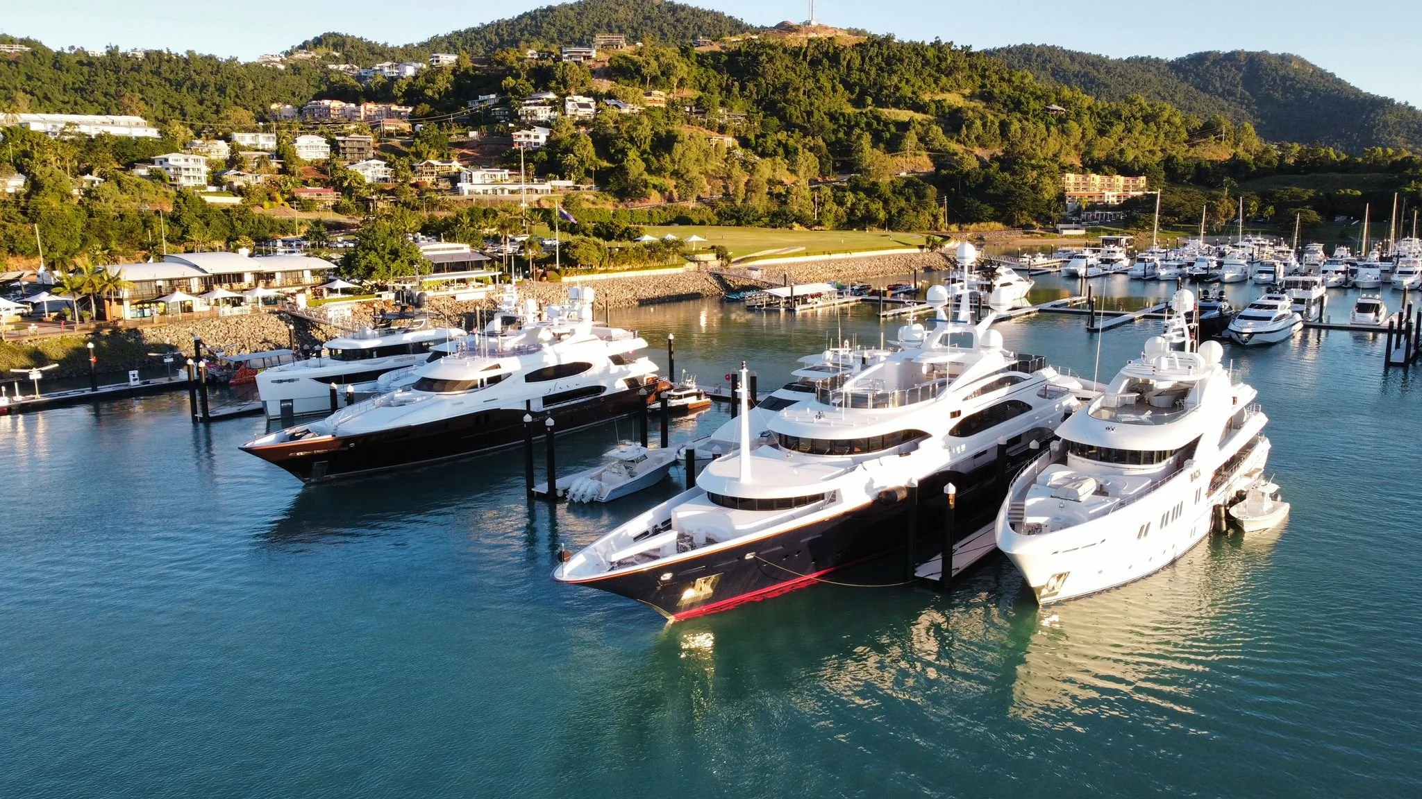 A marina with multiple large luxury yachts docked in calm water, surrounded by lush green hills with houses on the hillside.