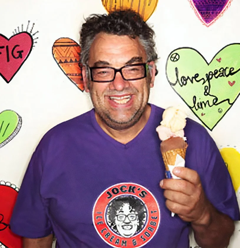 Jock Main standing in front of the hand painted wall inside Jock's Ice Cream and Sorbet Albert Park, wearing a purple jocks t-shirts and holding an icecream cone.