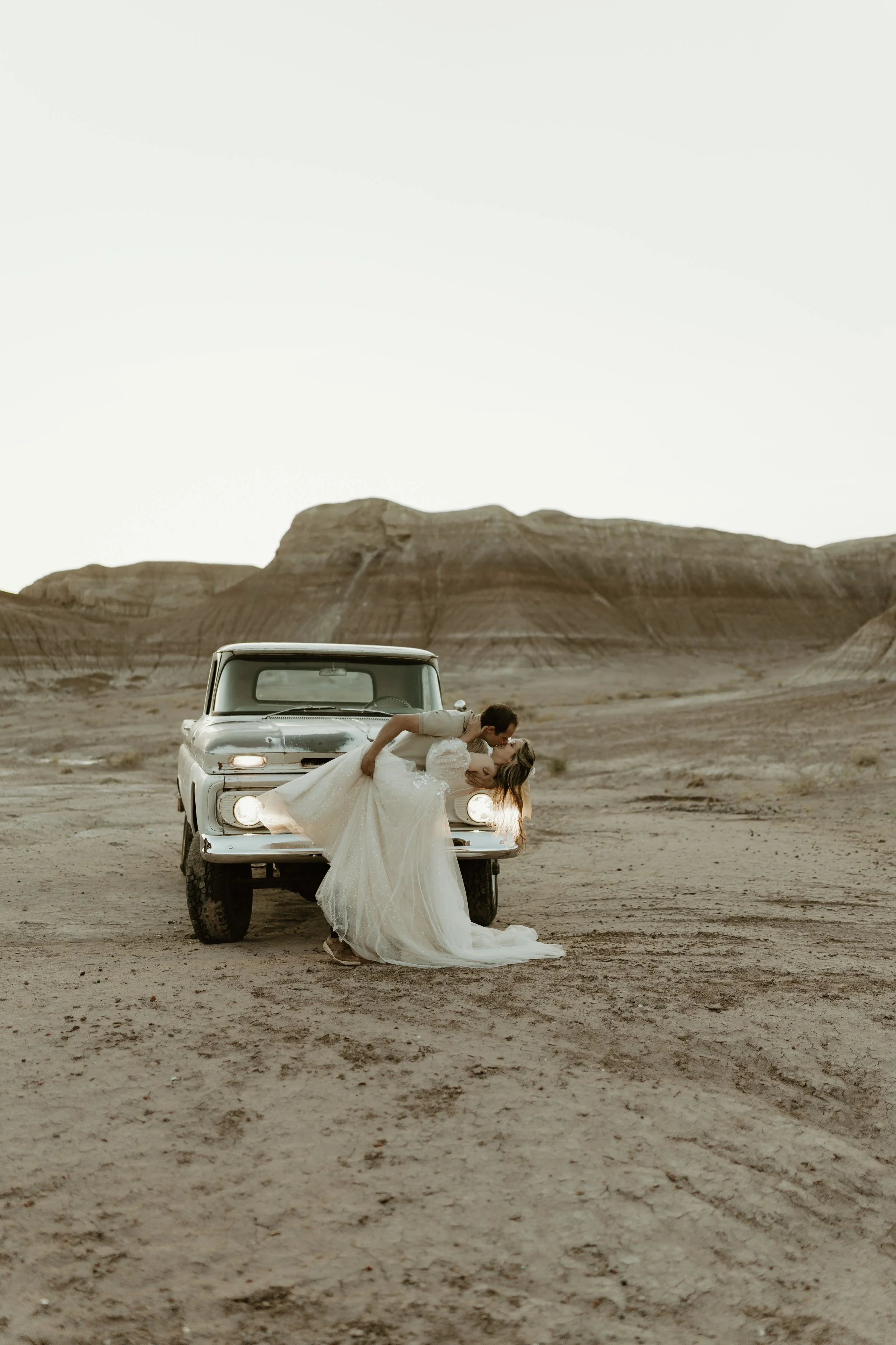 A newlywed couple sharing a kiss on the front of a vintage car in a desert landscape with large rock formations.