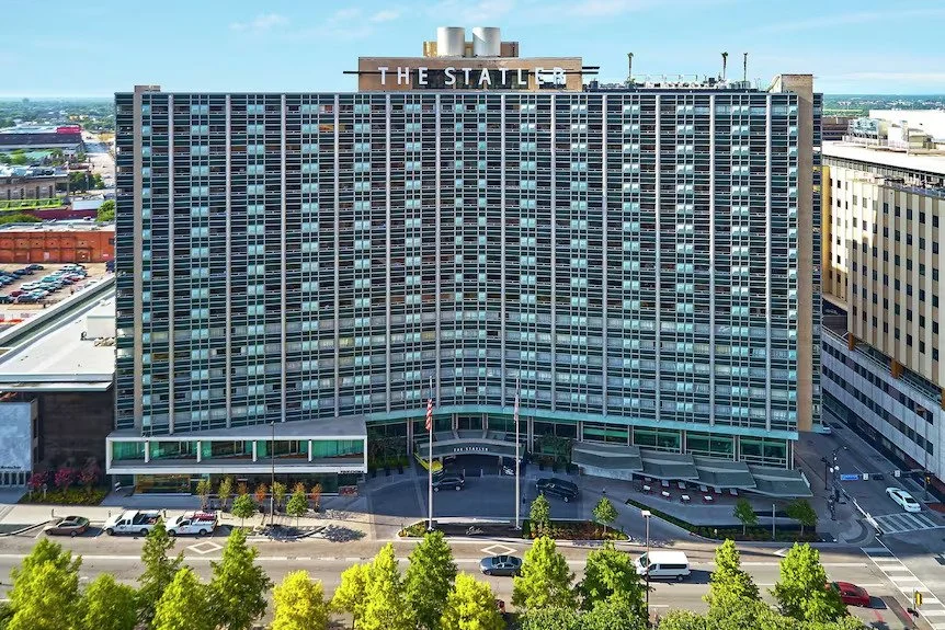 Tall modern hotel building with a curved facade and the sign 'The Starler' on top, located on a city street with trees and parked cars in front.