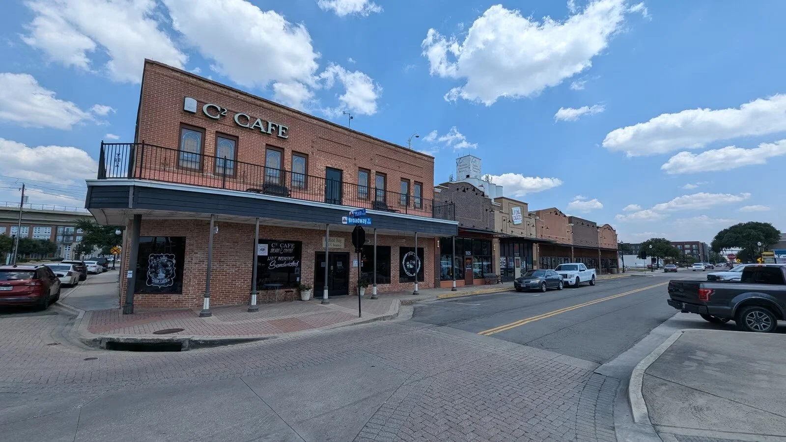 A two-story brick building with signs reading 'C2 CAFE' and a street sign at the corner indicating 'Broadway'. The building has a balcony and large windows, with a parking lot and cars in front. The sky is partly cloudy with blue sky visible.