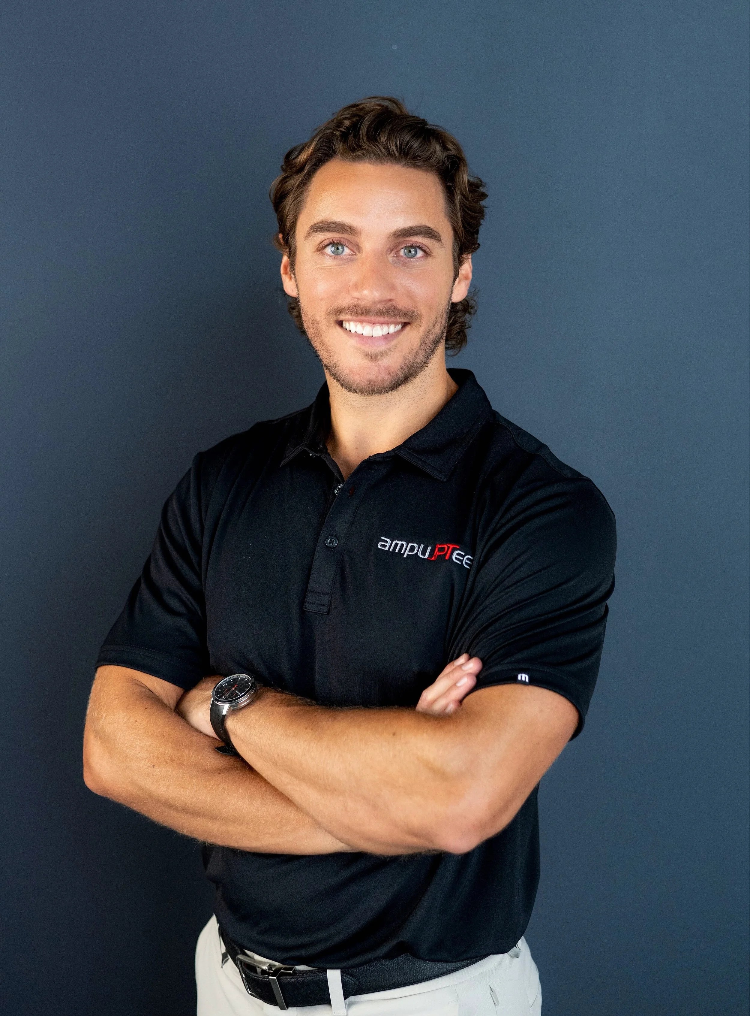 A young man with curly brown hair, blue eyes, and a beard, smiling and standing with arms crossed, wearing a black polo shirt with a logo, against a dark gray background.