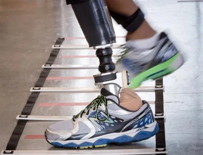 A prosthetic leg is being used to kick a running shoe off a foot, with a treadmill or workout area in the background.