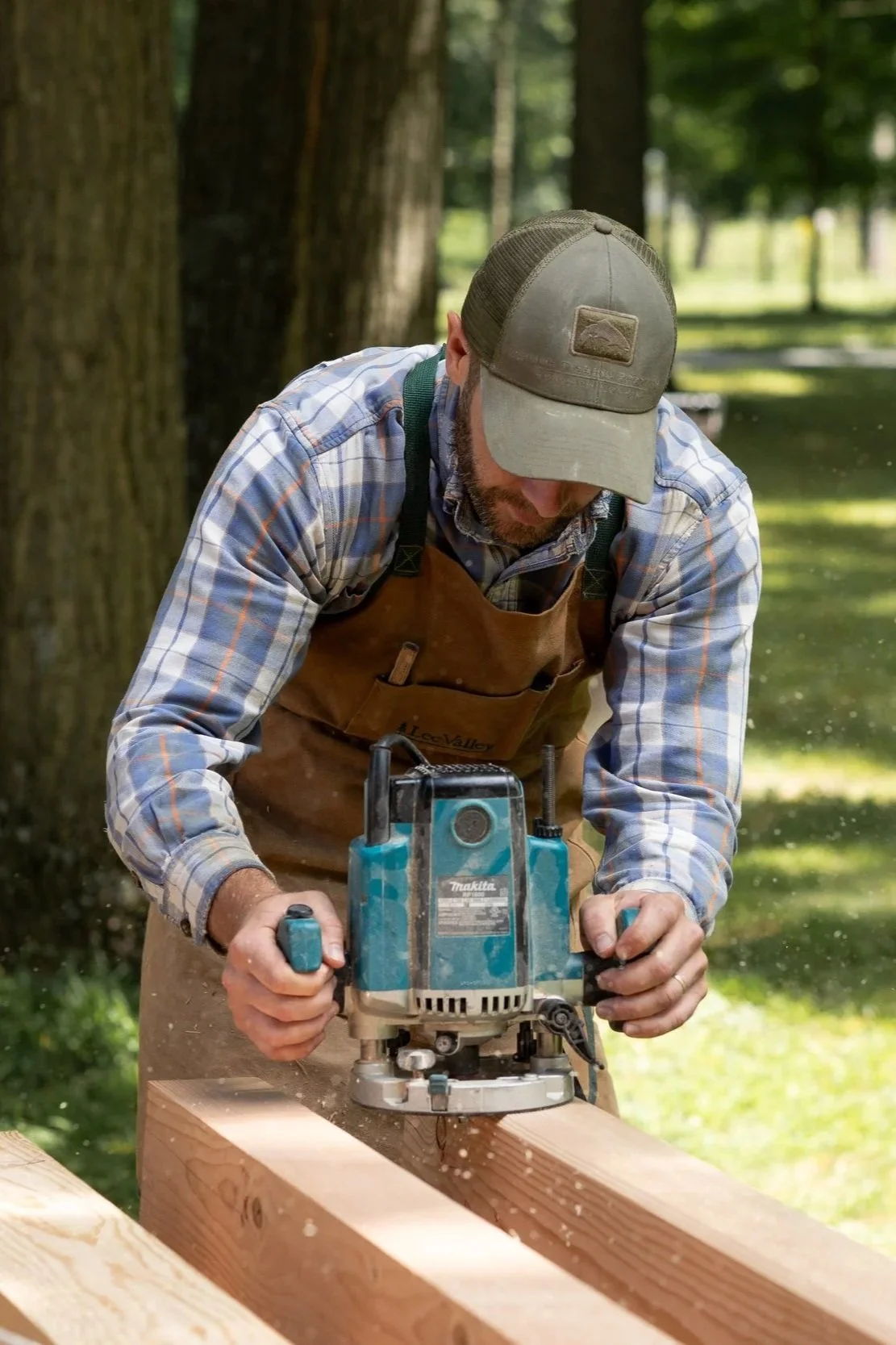 A man wearing a cap, plaid shirt, and apron using a circular saw on a wooden beam outdoors among trees.